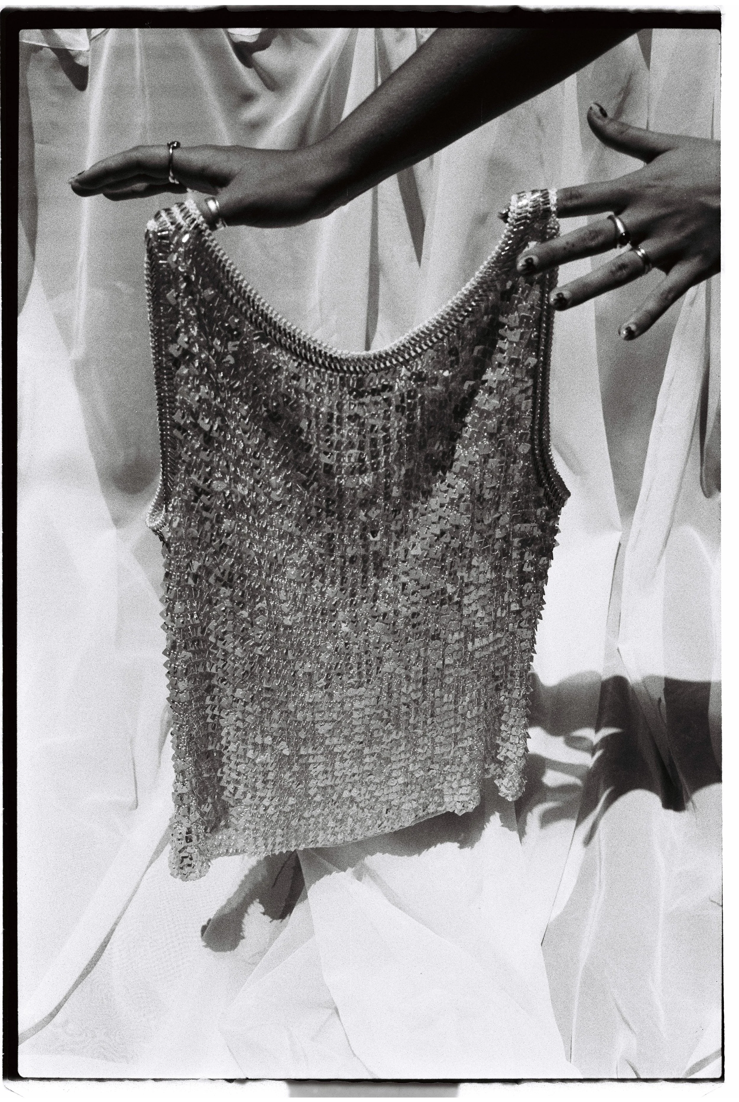 A person holding a shiny, textured handbag with metallic details, wearing multiple rings on their fingers, the background appears to be crumpled paper or fabric.
