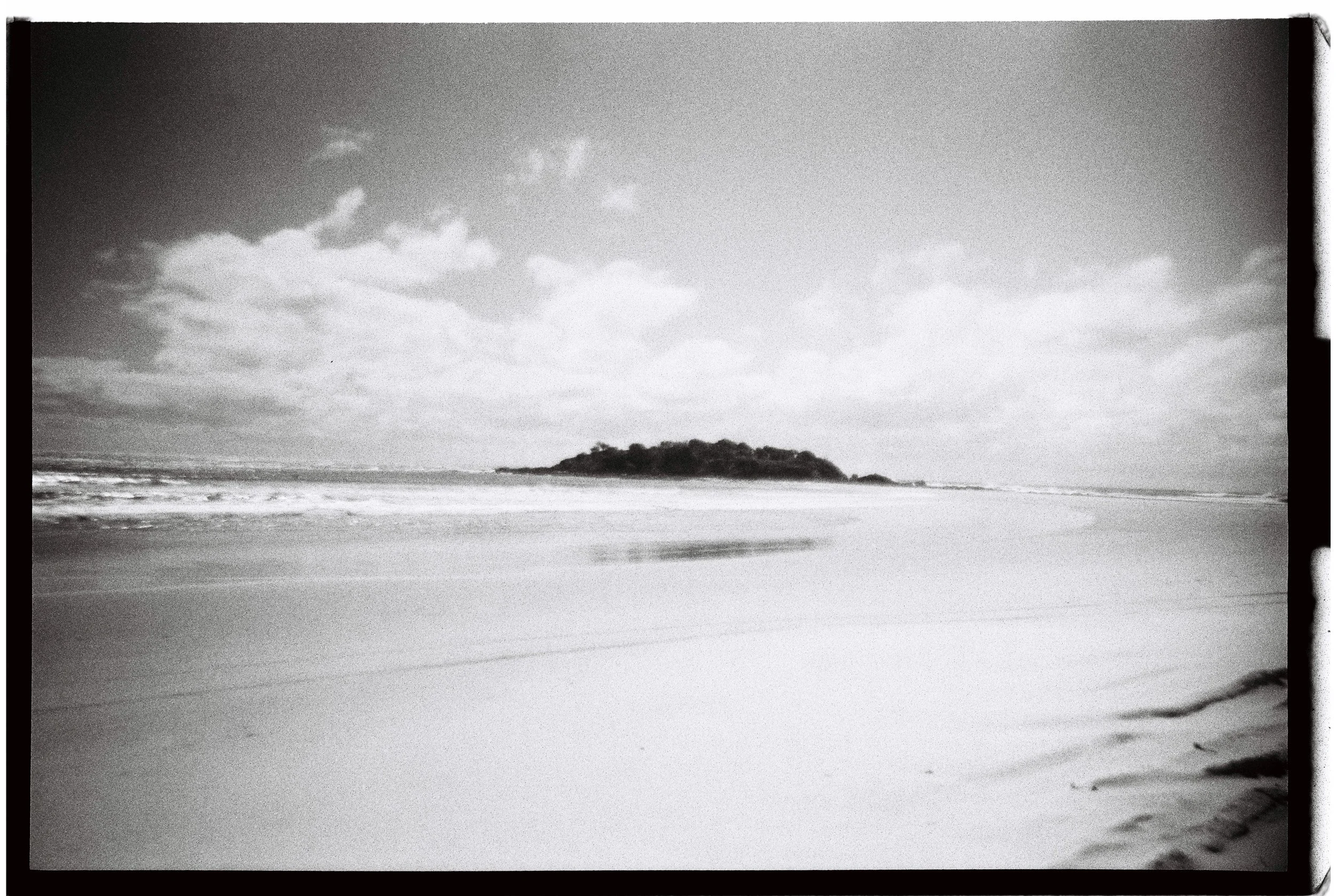 Black and white photo of a beach with an island in the distance, cloudy sky, waves crashing on the shore.