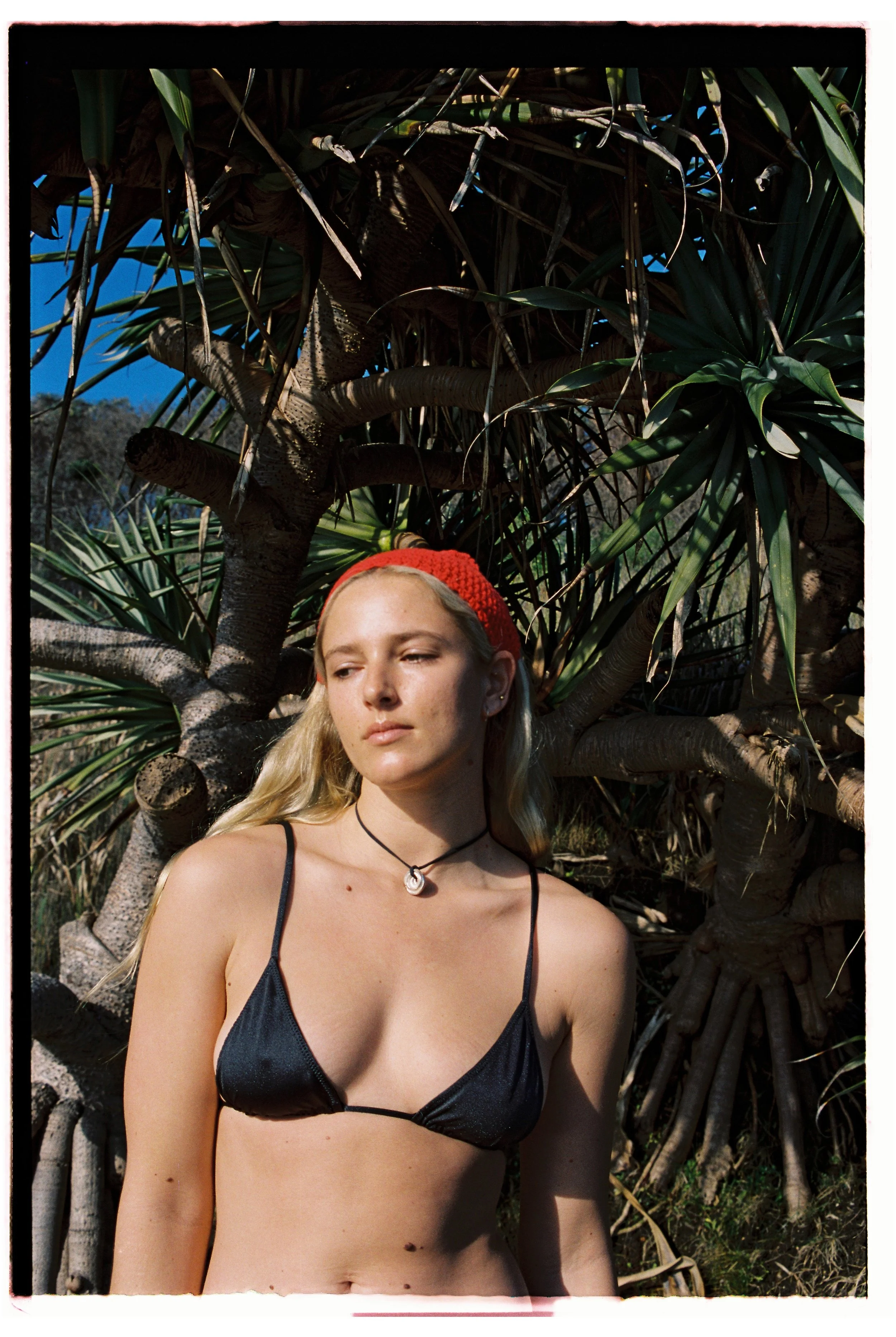 A young woman in a black bikini top and a red headband standing outdoors with large leafy plants and a blue sky in the background.