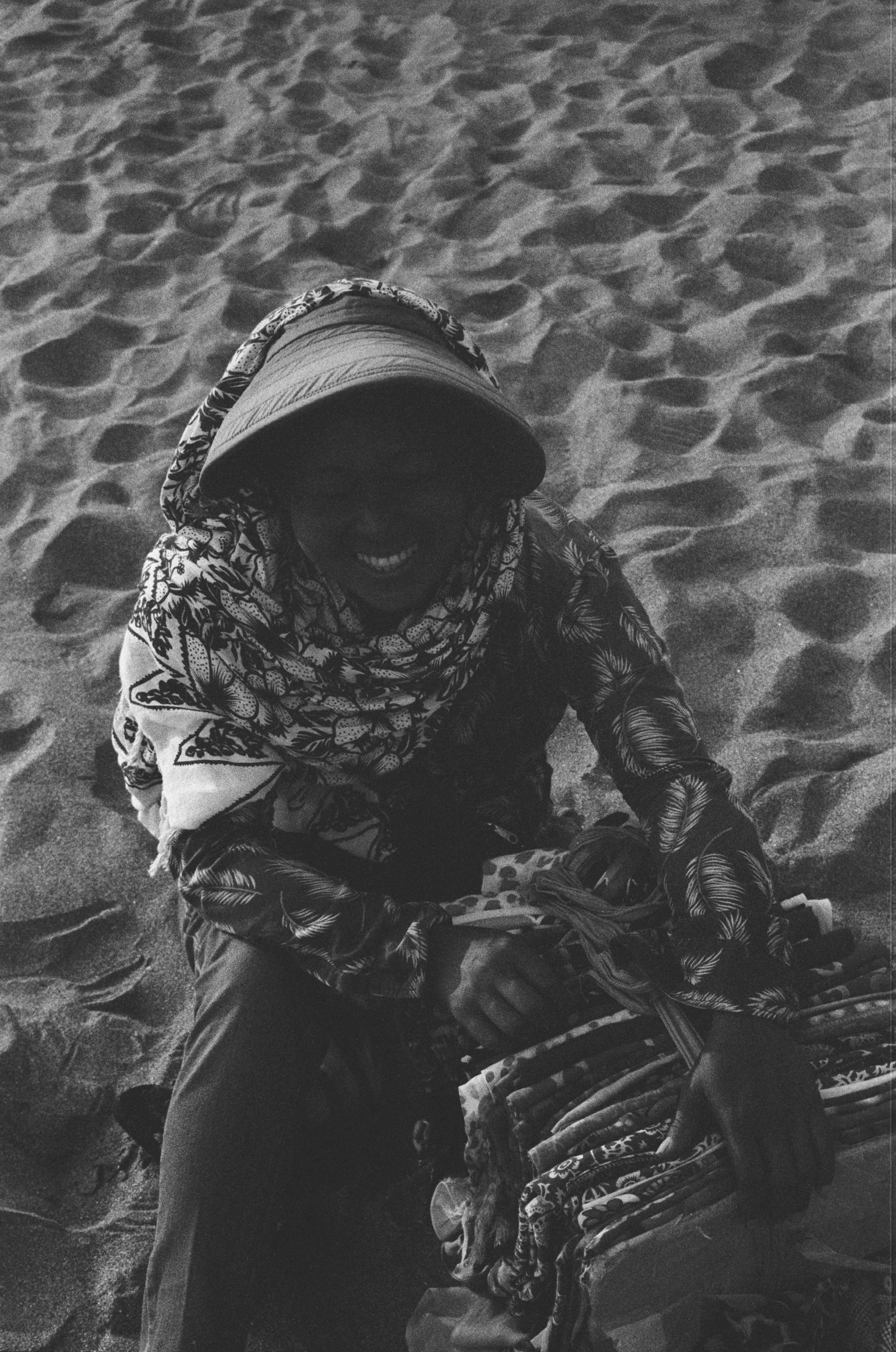 Person sitting on sandy beach, wearing a cap, patterned scarf, and gloves, smiling, with supplies or clothing in front.