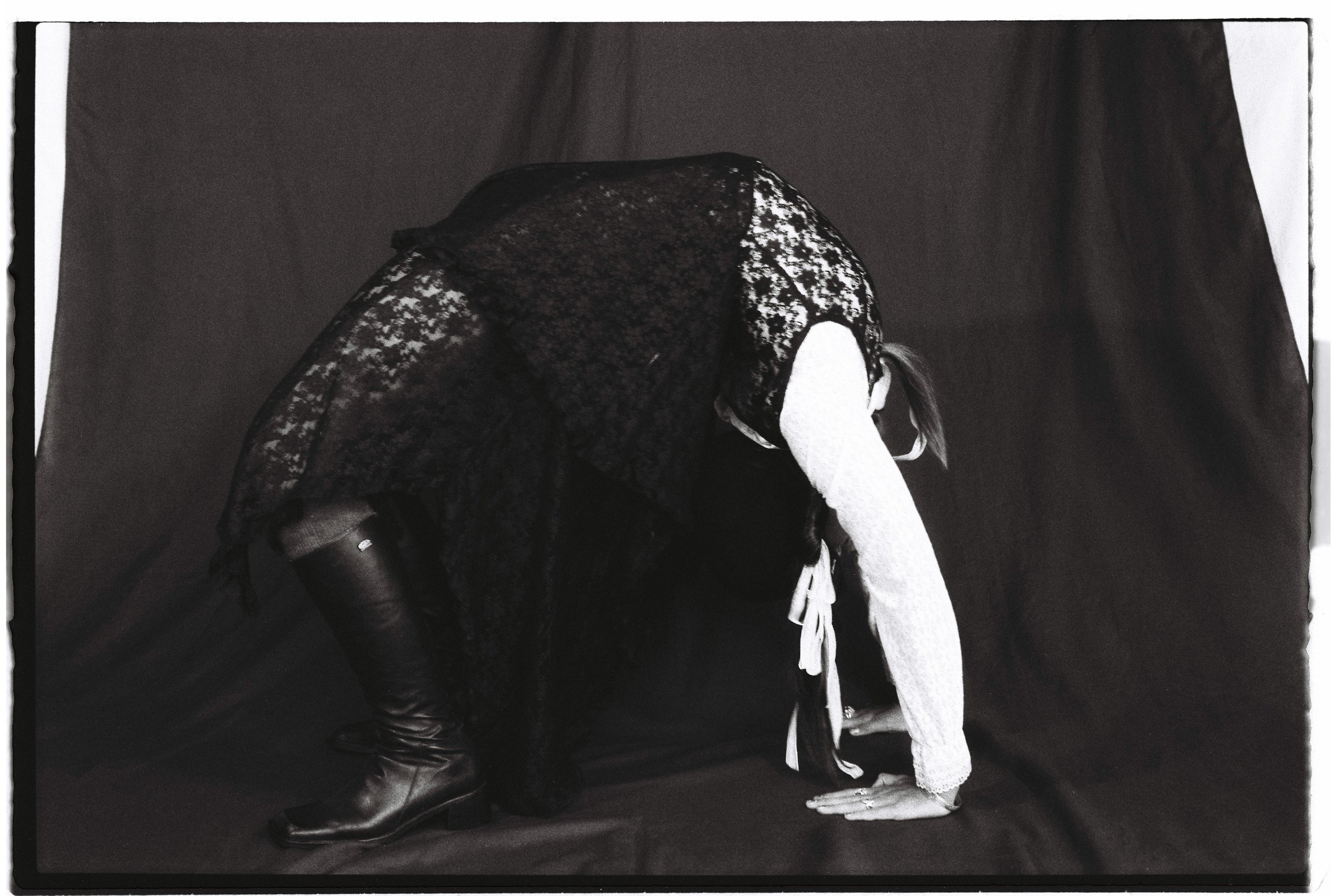 A woman dressed in a lace dress and leather boots poses on a black backdrop, leaning forward with her hands on the ground and her head bowed.