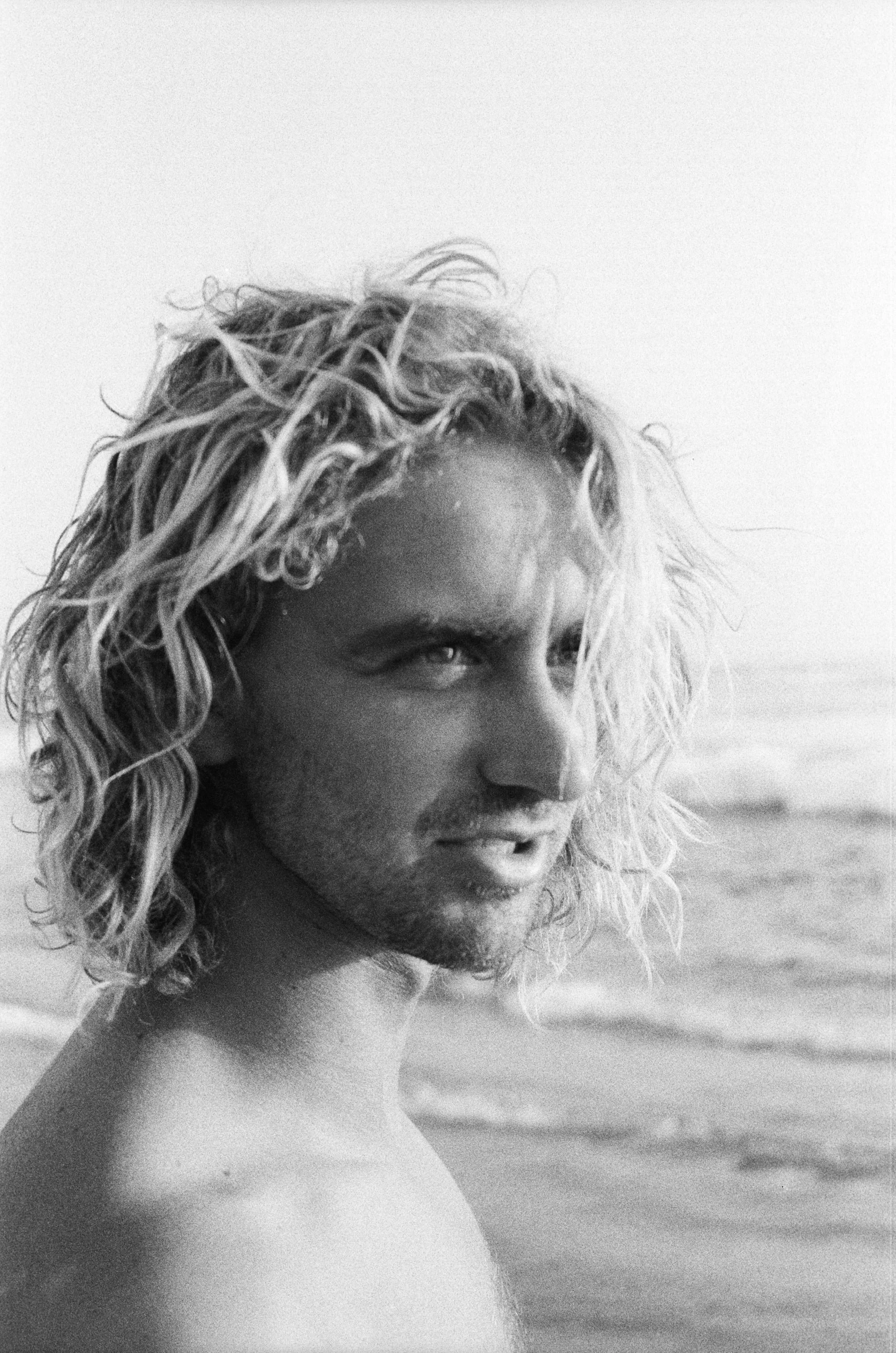A black and white photograph of a person with long, curly hair at the beach, looking toward the camera with a slight smile.
