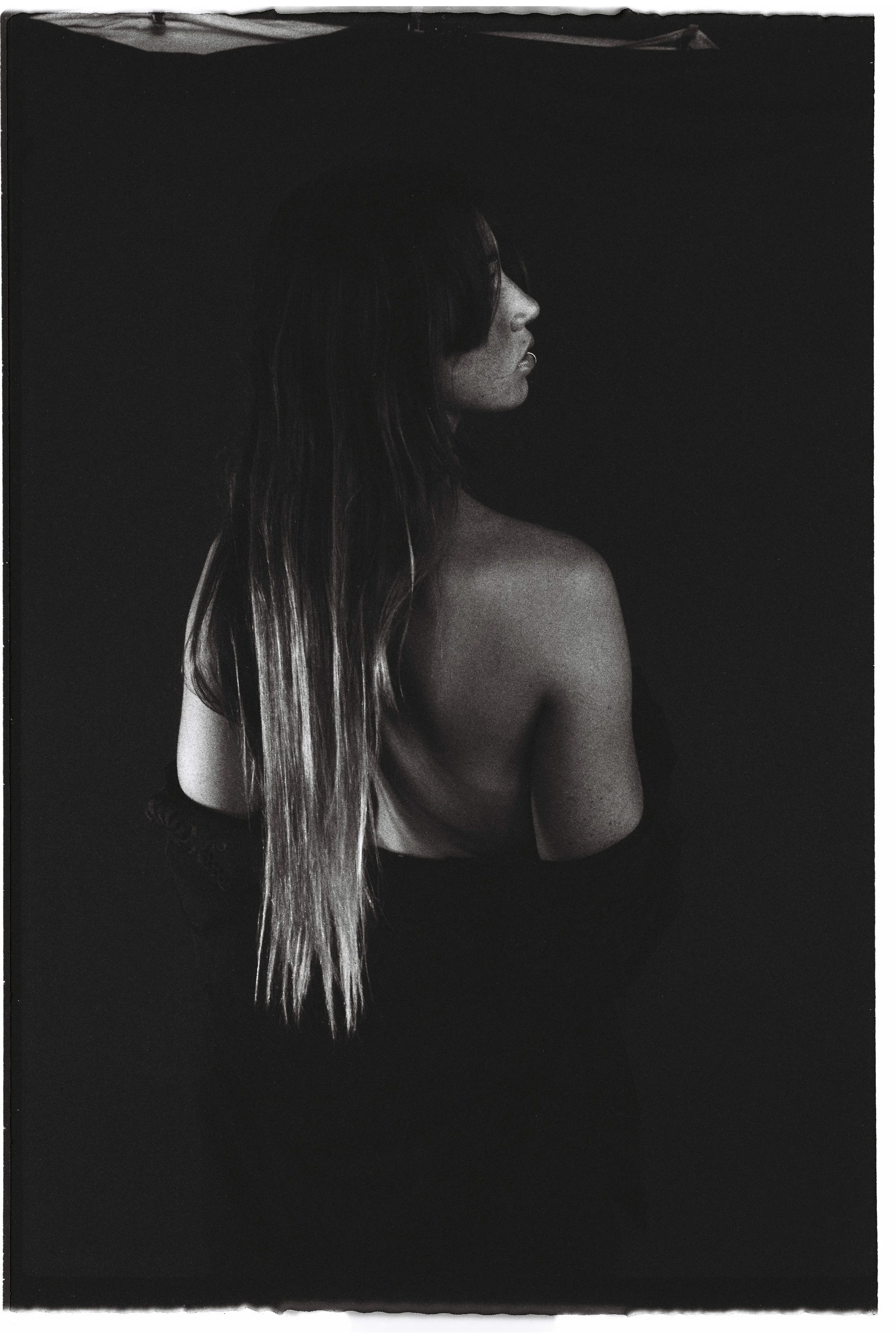 Black and white photo of a woman with long hair, turned to the side, with her back partially exposed, against a dark background.