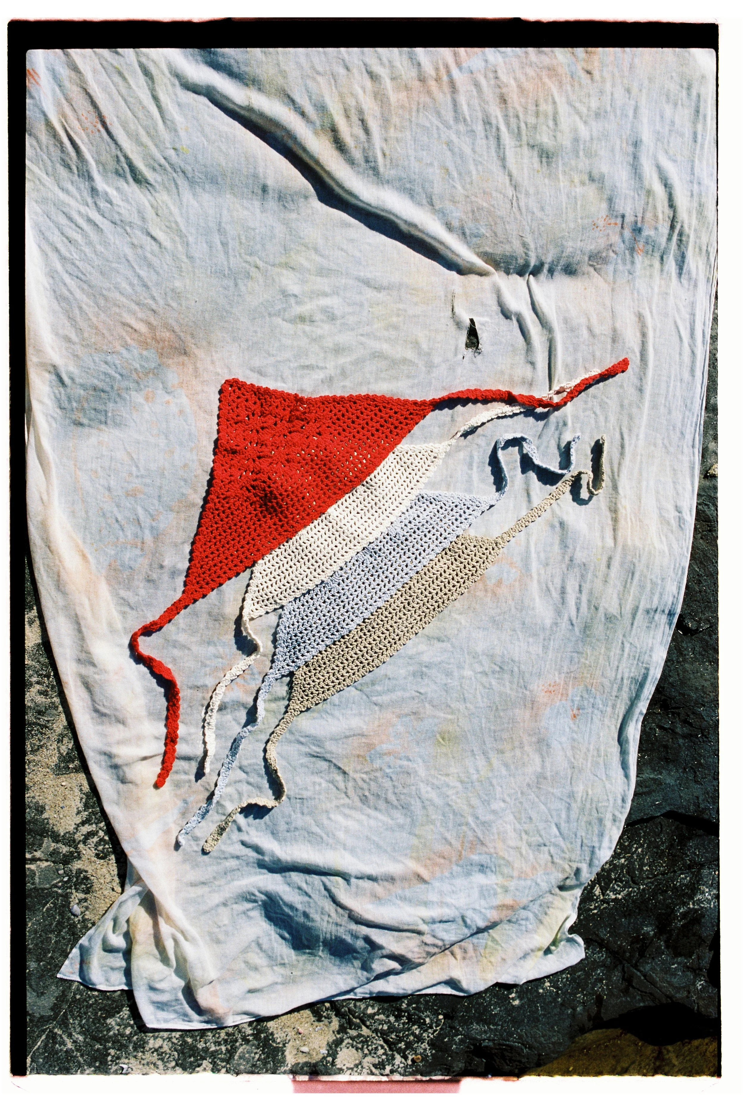 A white cloth laid on a surface with four colorful crocheted manta ray-shaped pieces in red, white, light blue, and beige.