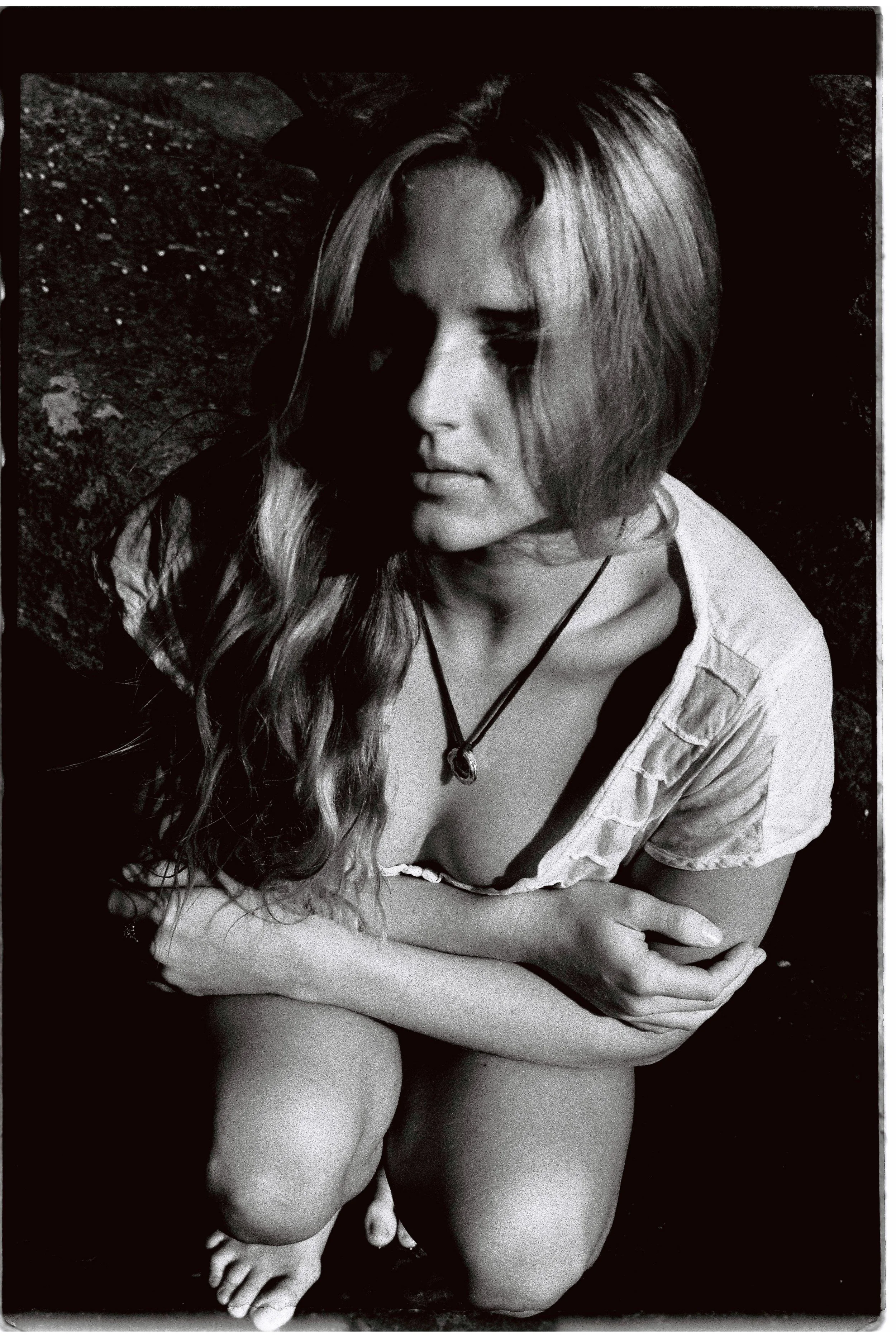 A young woman with long wavy hair, wearing a light-colored shirt and a necklace, is sitting outdoors on her knees with her arms crossed around her knees, in black and white.