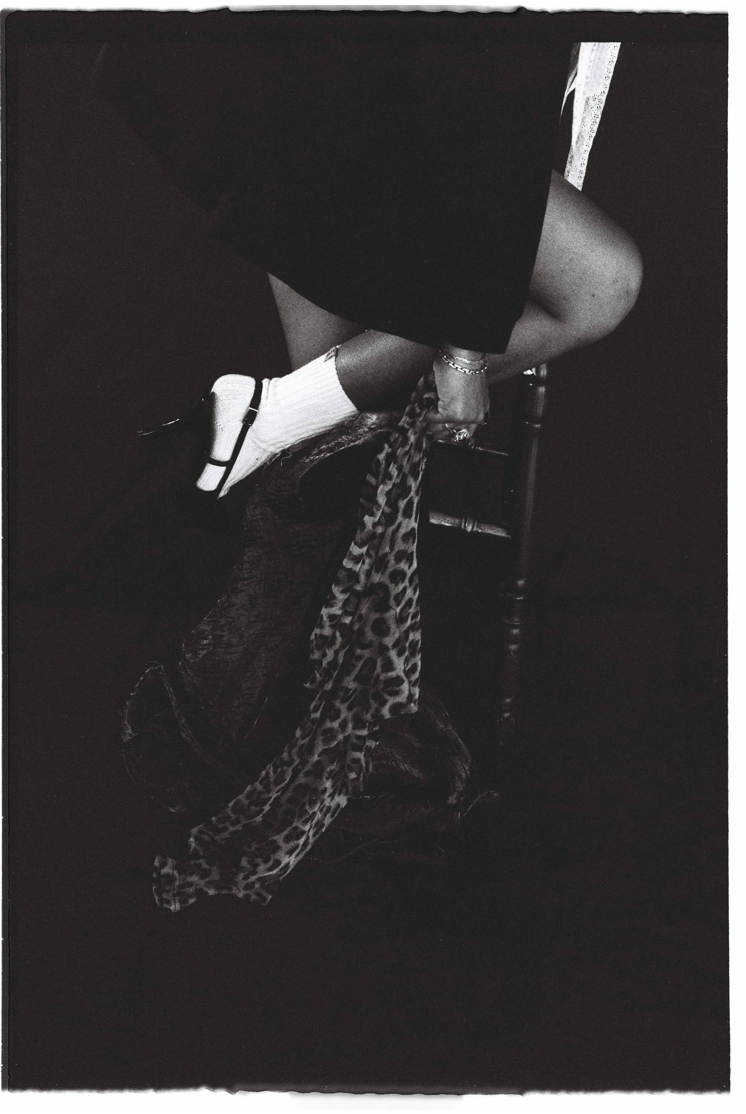 A person sitting on a wooden chair with one leg crossed over the other. They are wearing a dark skirt, white socks, and black sandals. Their hand, with rings and bracelets, is resting on a leopard print cloth hanging from the chair. The background is