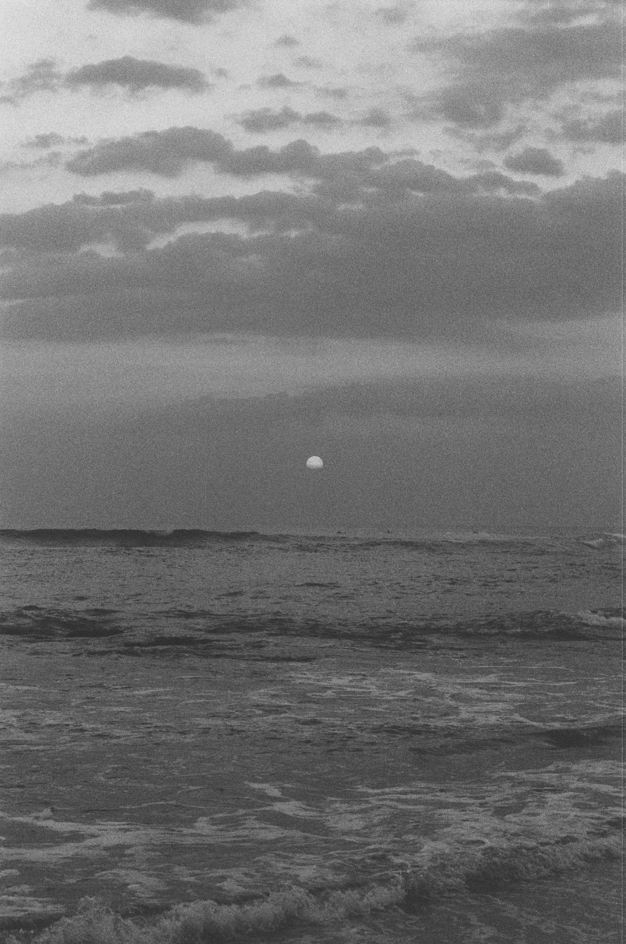 Black and white photo of the ocean with waves, clouds in the sky, and the moon near the horizon.