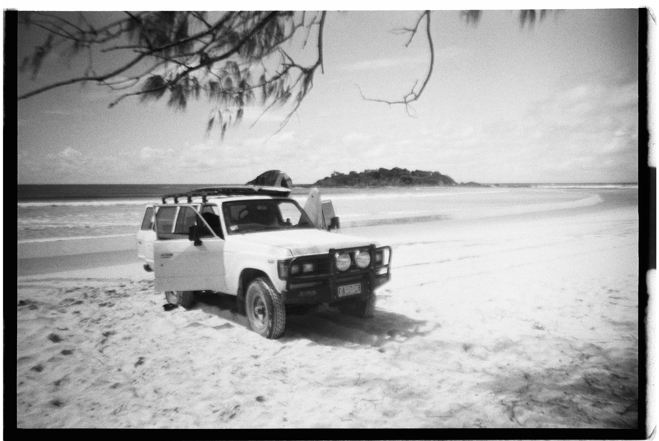 A black and white photo of a car parked on a sandy beach, with the ocean and an island in the background, and a tree branch overhead.