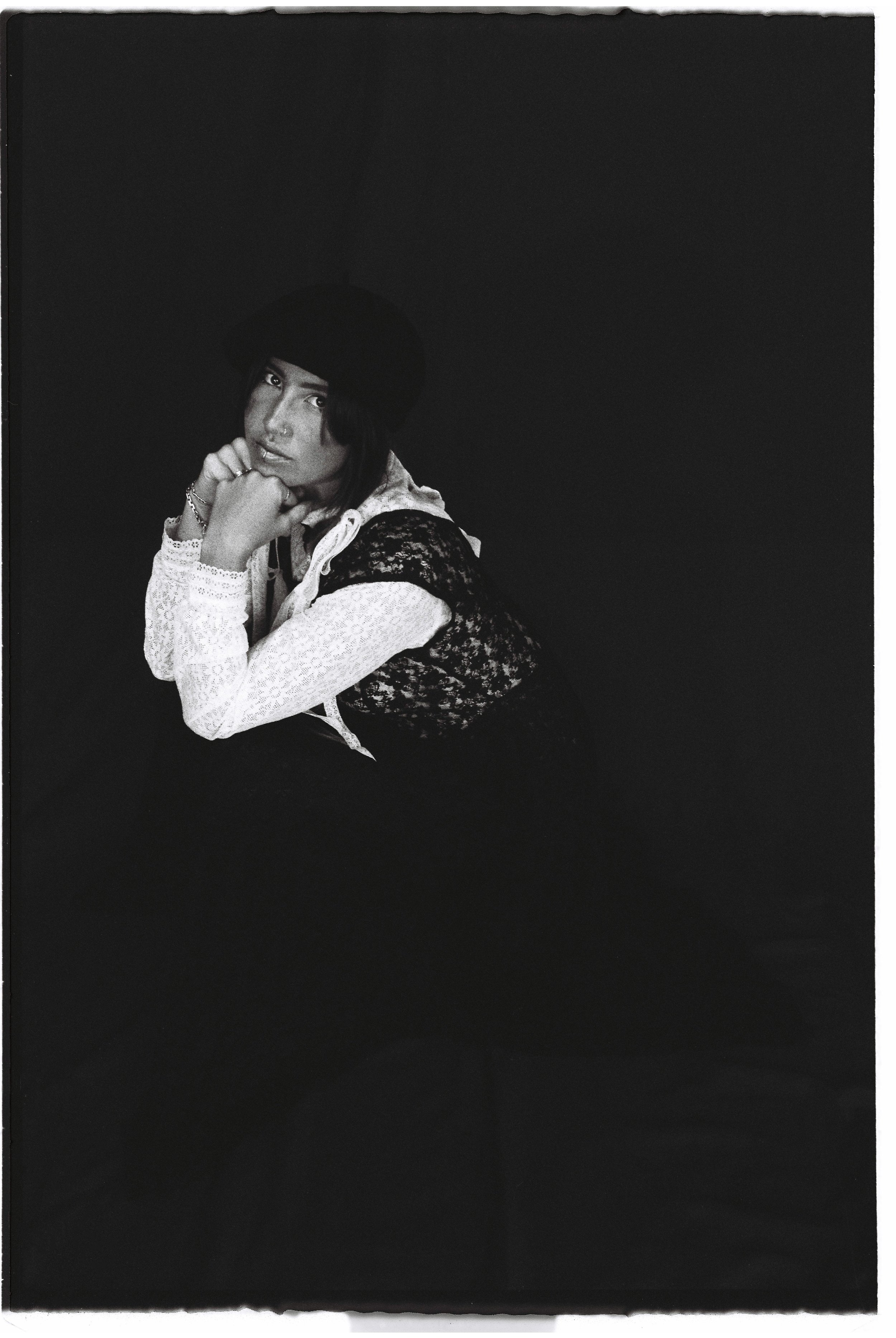 A person with long hair wearing a hat, lace top, and a hoodie, resting their chin on folded arms against a dark background.