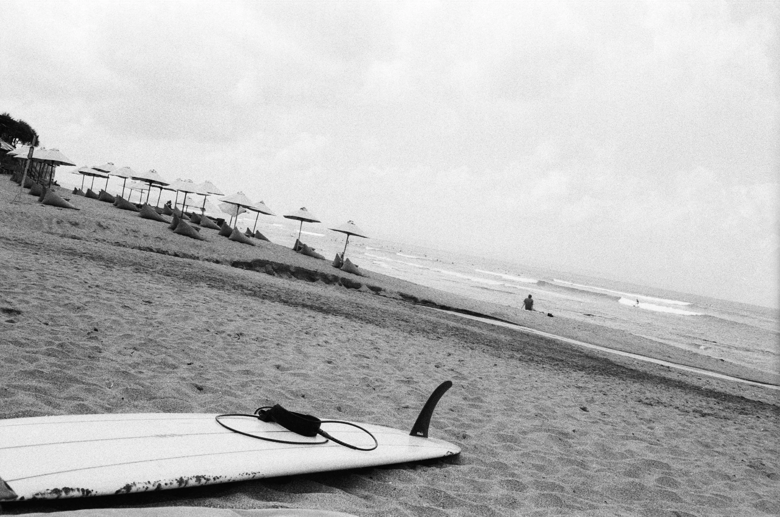 Black and white photo of a surfboard on the sand near the shoreline with a leash attached, empty beach with umbrellas in the background, a person sitting on the sand near the water, and waves in the ocean.