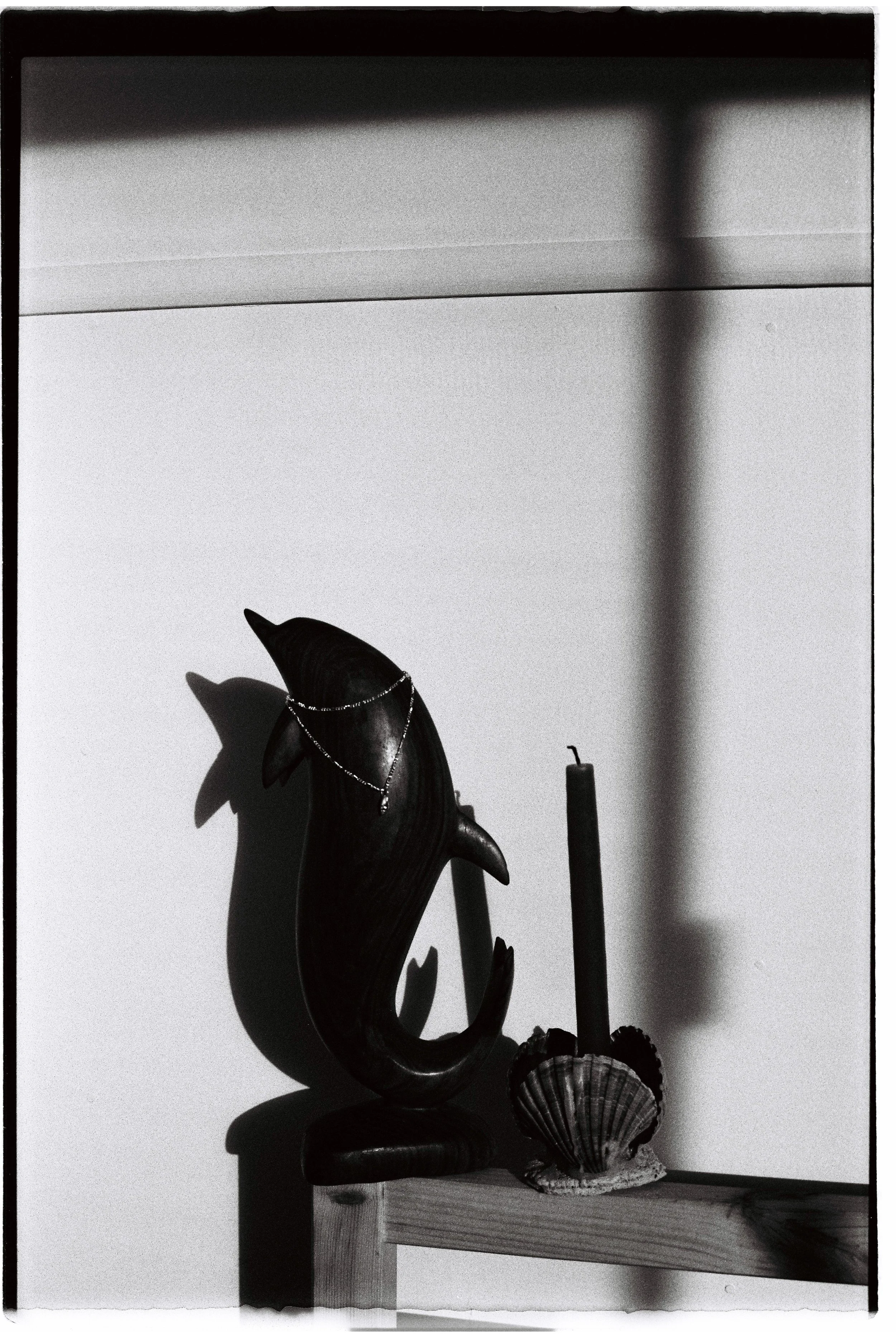 A carved wooden dolphin sculpture with a necklace, a candle, and a seashell on a wooden shelf, casting shadows on the wall.