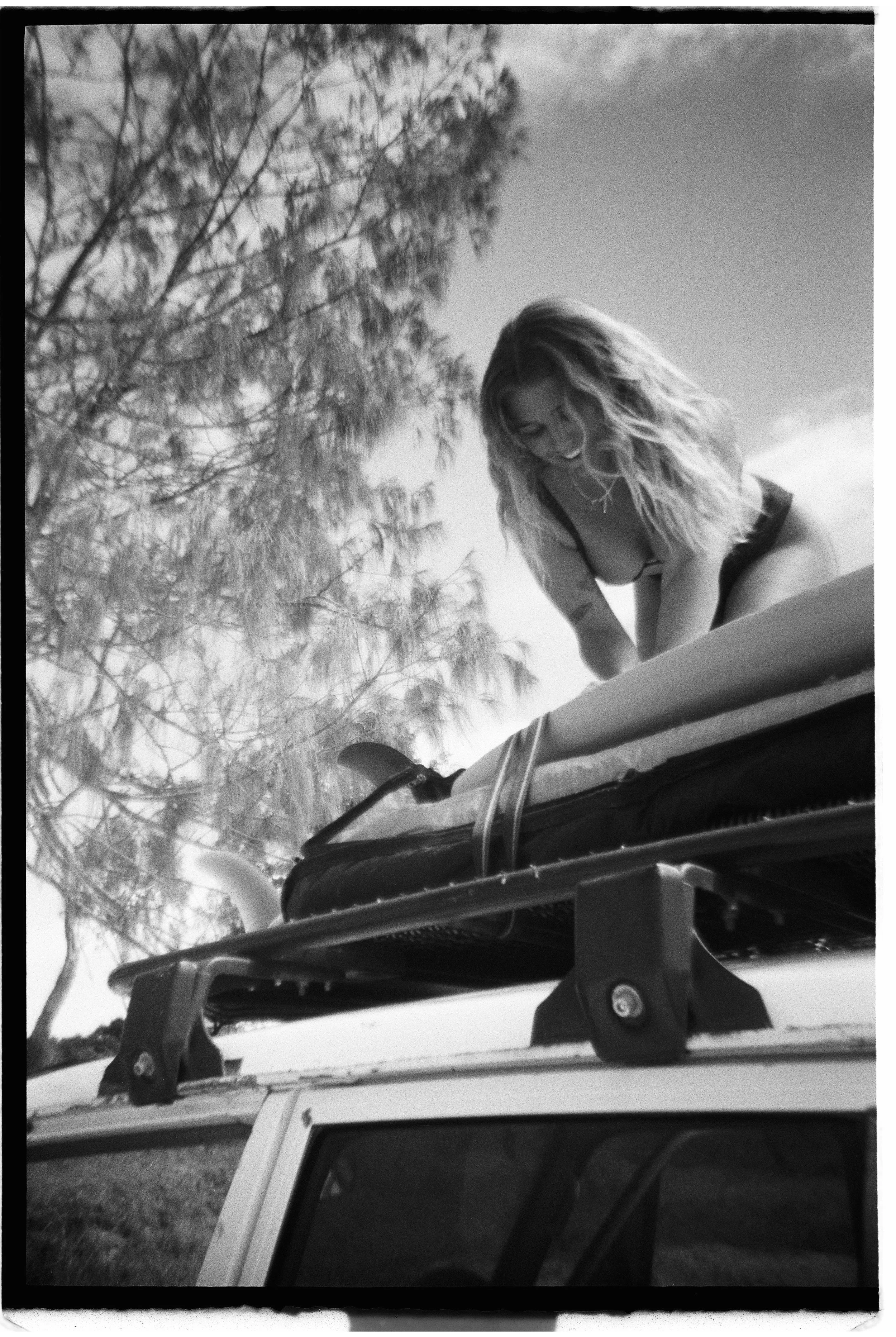 A woman with long, wavy hair leaning over the roof of a vehicle, smiling, with background trees and a clear sky.