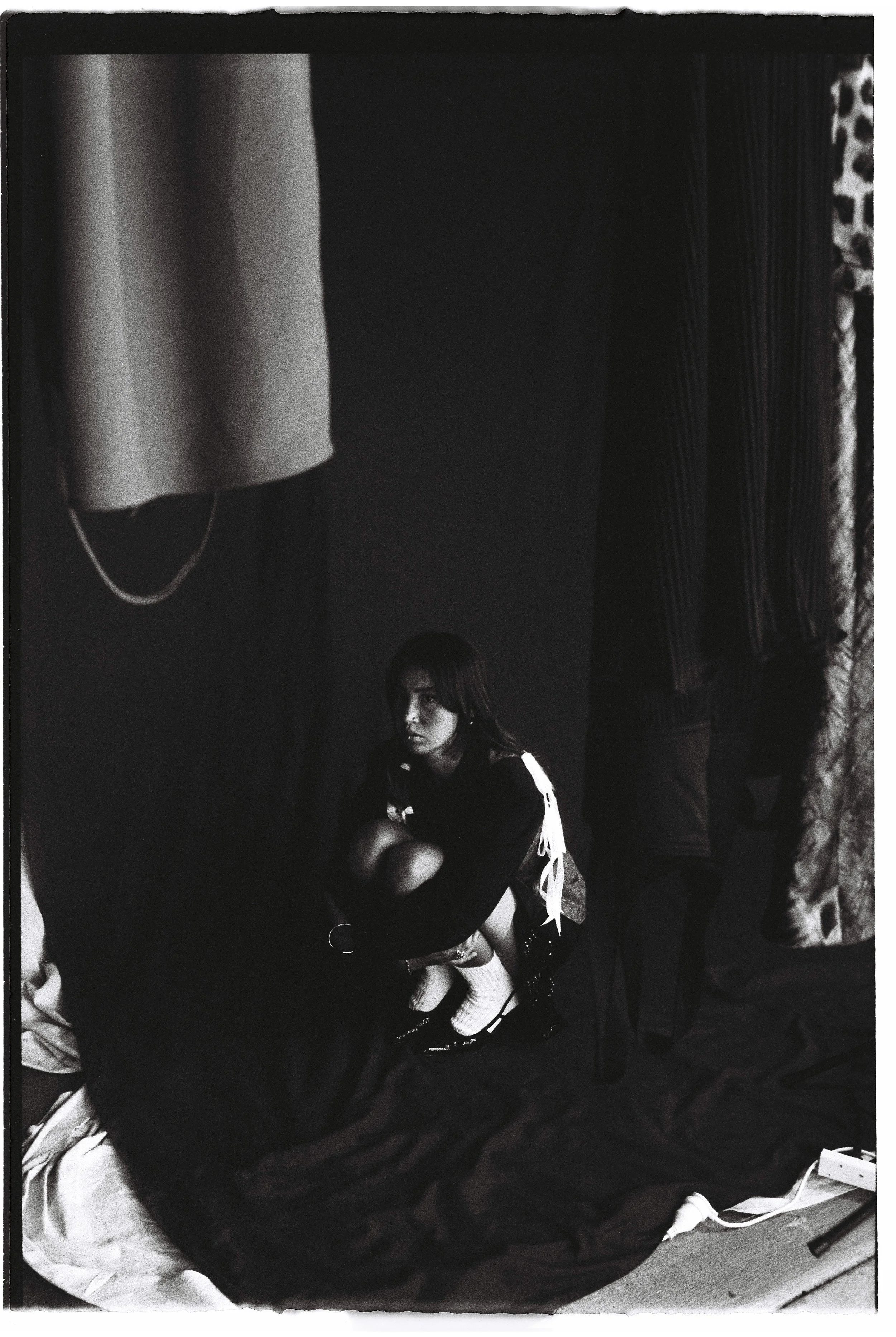 A woman crouched on the floor in a dark room, with a curtain on the right and a large cloth hanging from the ceiling on the left, surrounded by dark fabric and some objects.