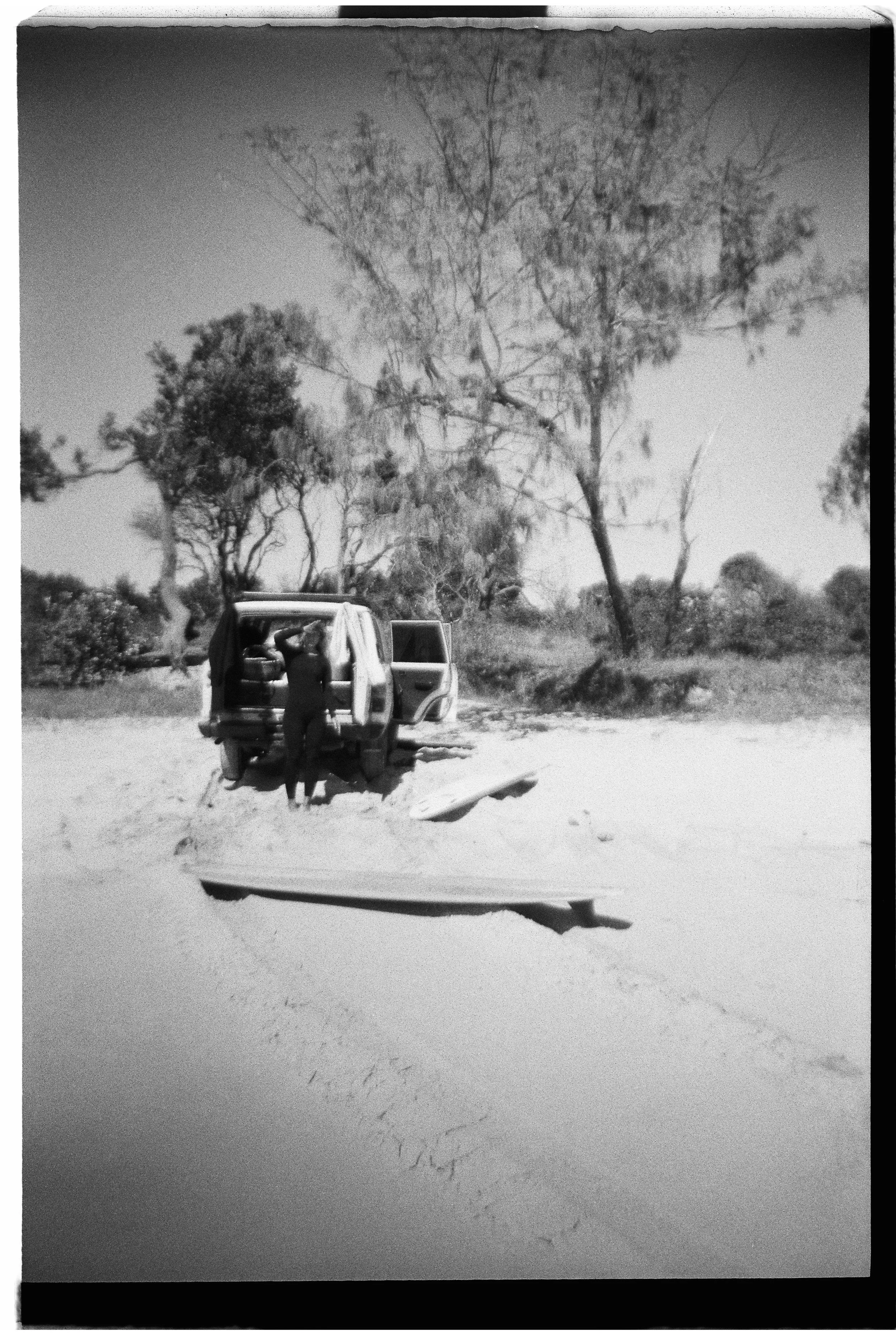 A person standing by an open vehicle door on a sandy landscape with trees in the background.