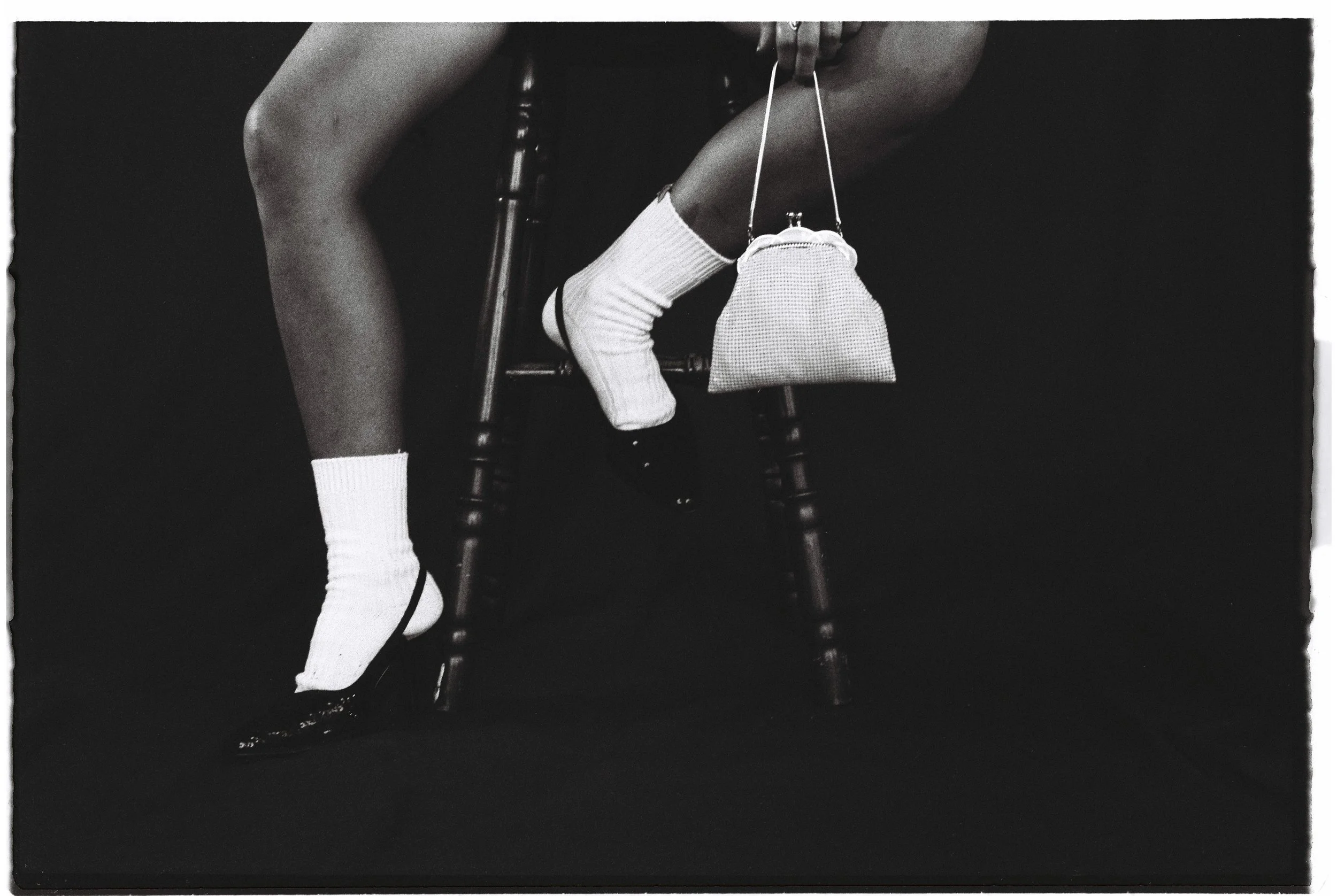 Black and white photo of a person's legs and feet sitting on a wooden chair, wearing white socks and shiny black shoes, with a small purse hanging from their ankle.
