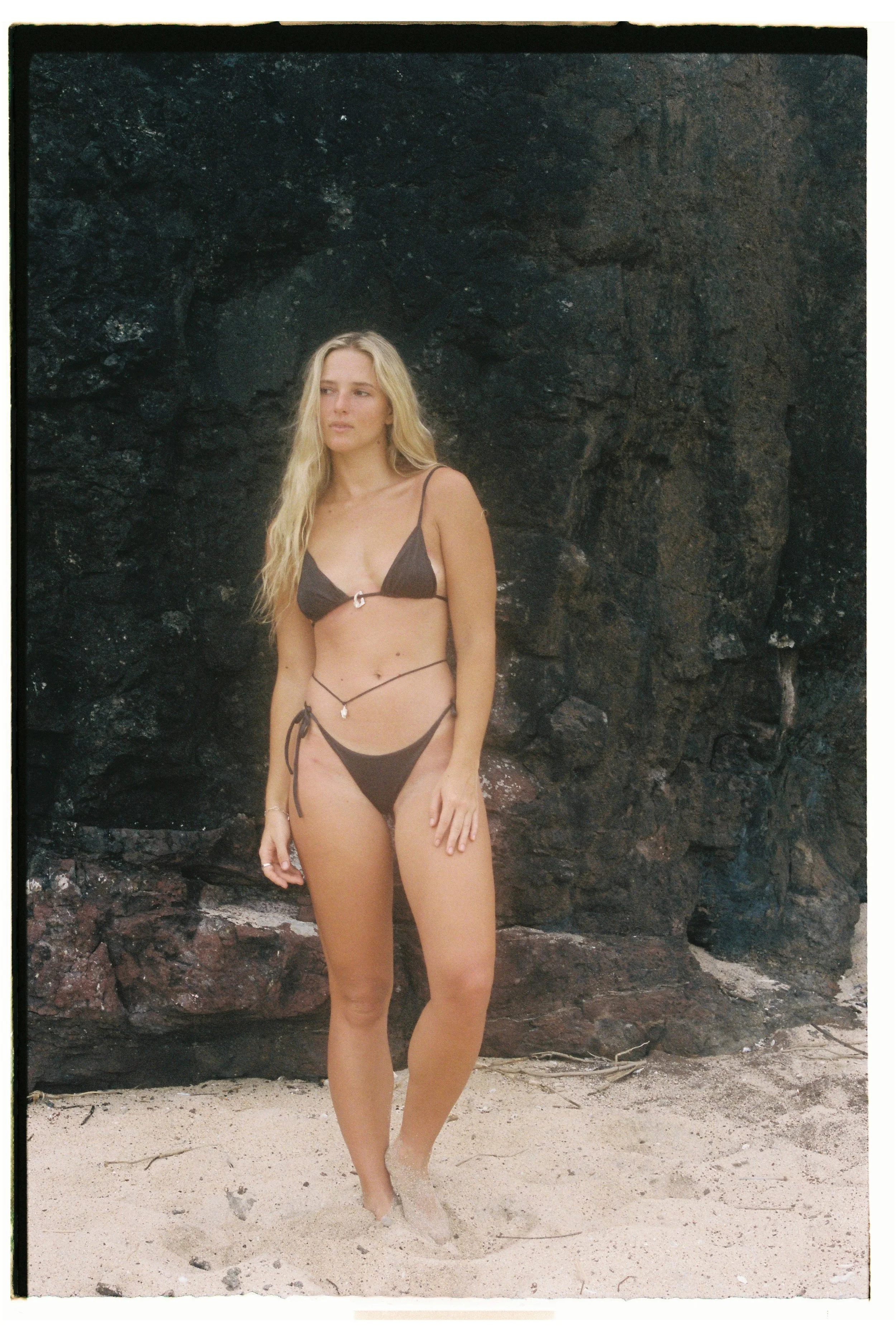 A woman wearing a dark bikini stands on sand in front of a dark rocky cliff.