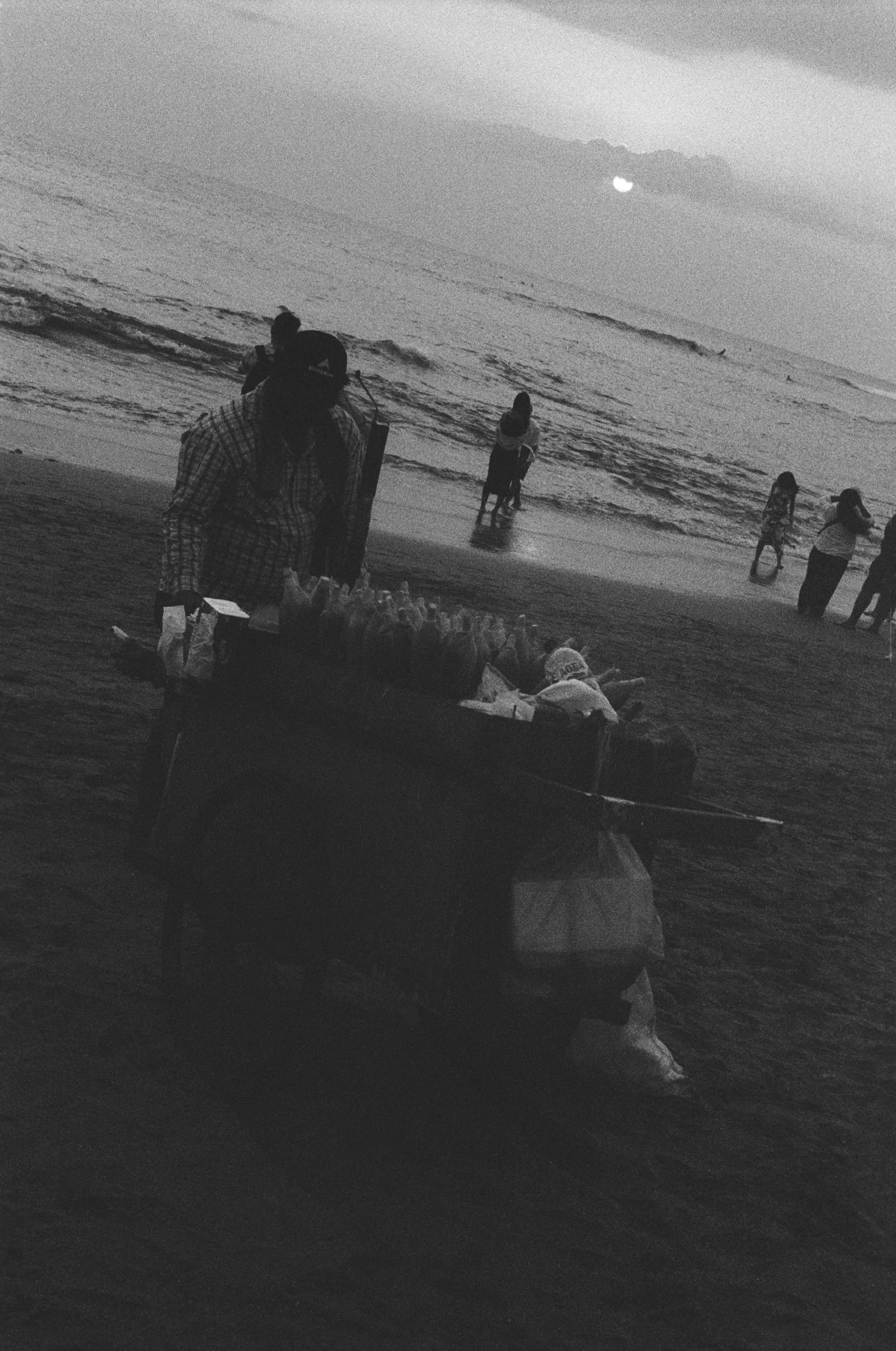 A person pushing a cart filled with items along a beach at sunset, with several people in the water and on the shore in the background.
