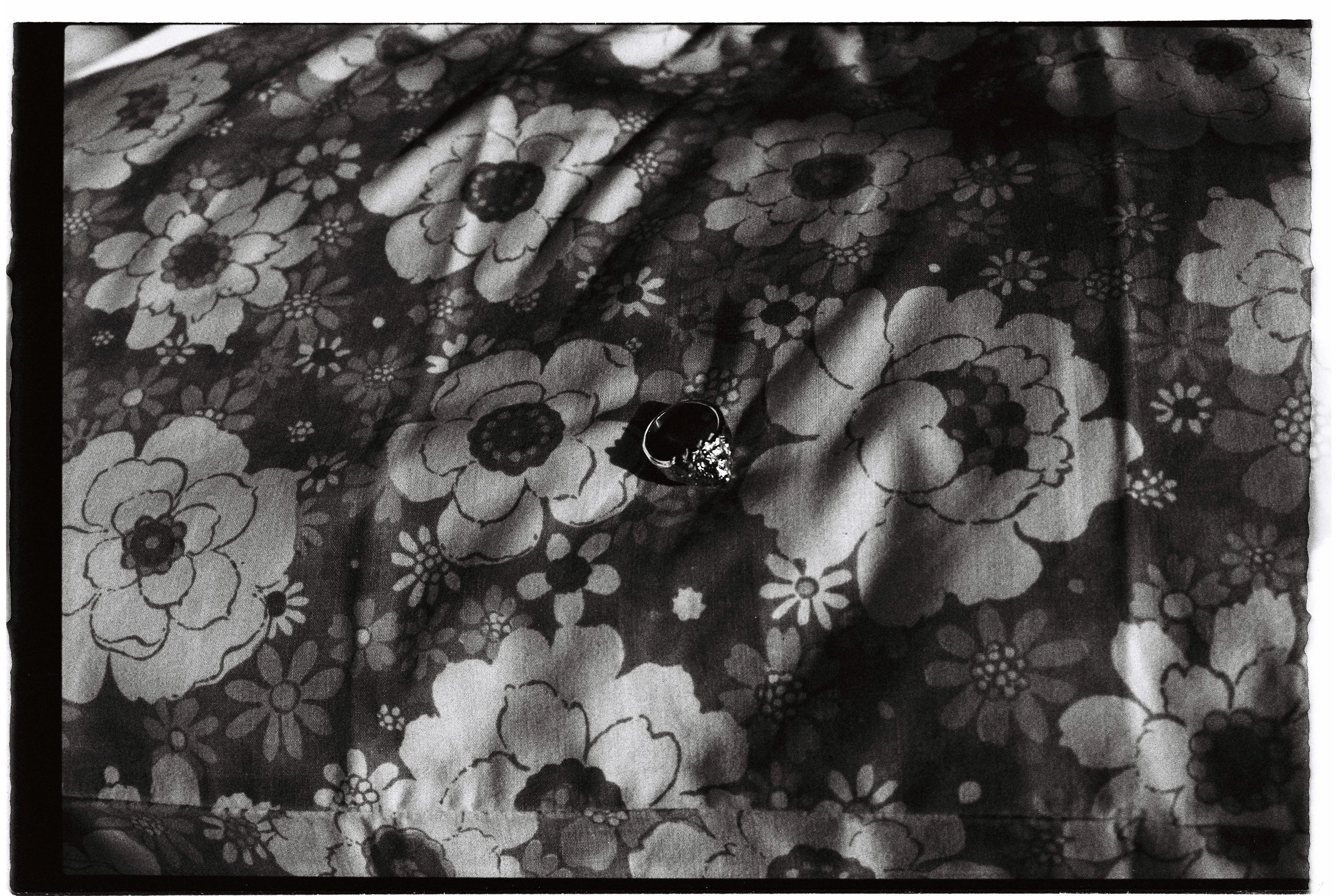 A close-up black and white photo of a patterned fabric with floral design, featuring a ring with a dark gemstone and metal band placed on it.