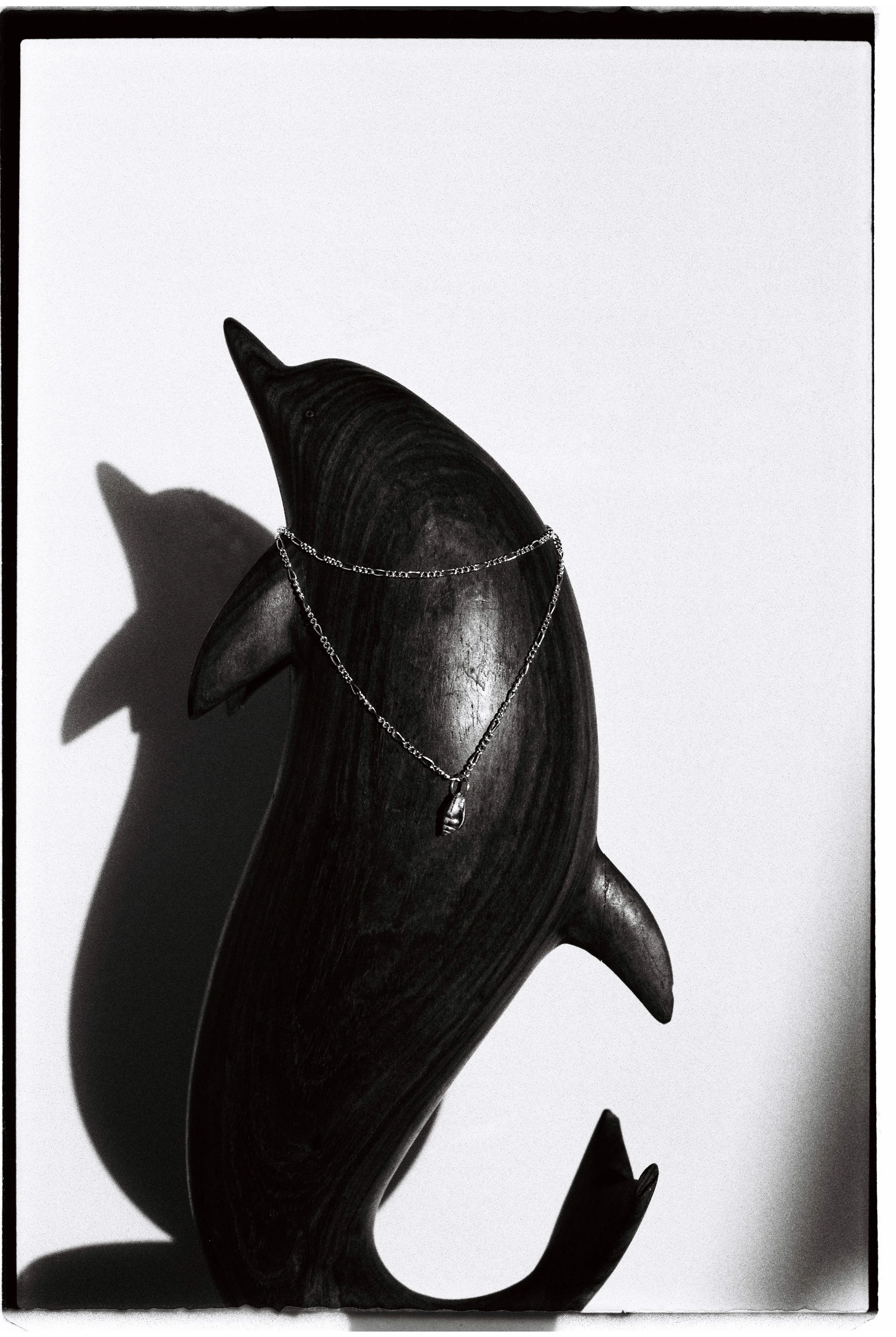 A wooden whale sculpture with a silver chain necklace hanging on it, casting a shadow on a white background.