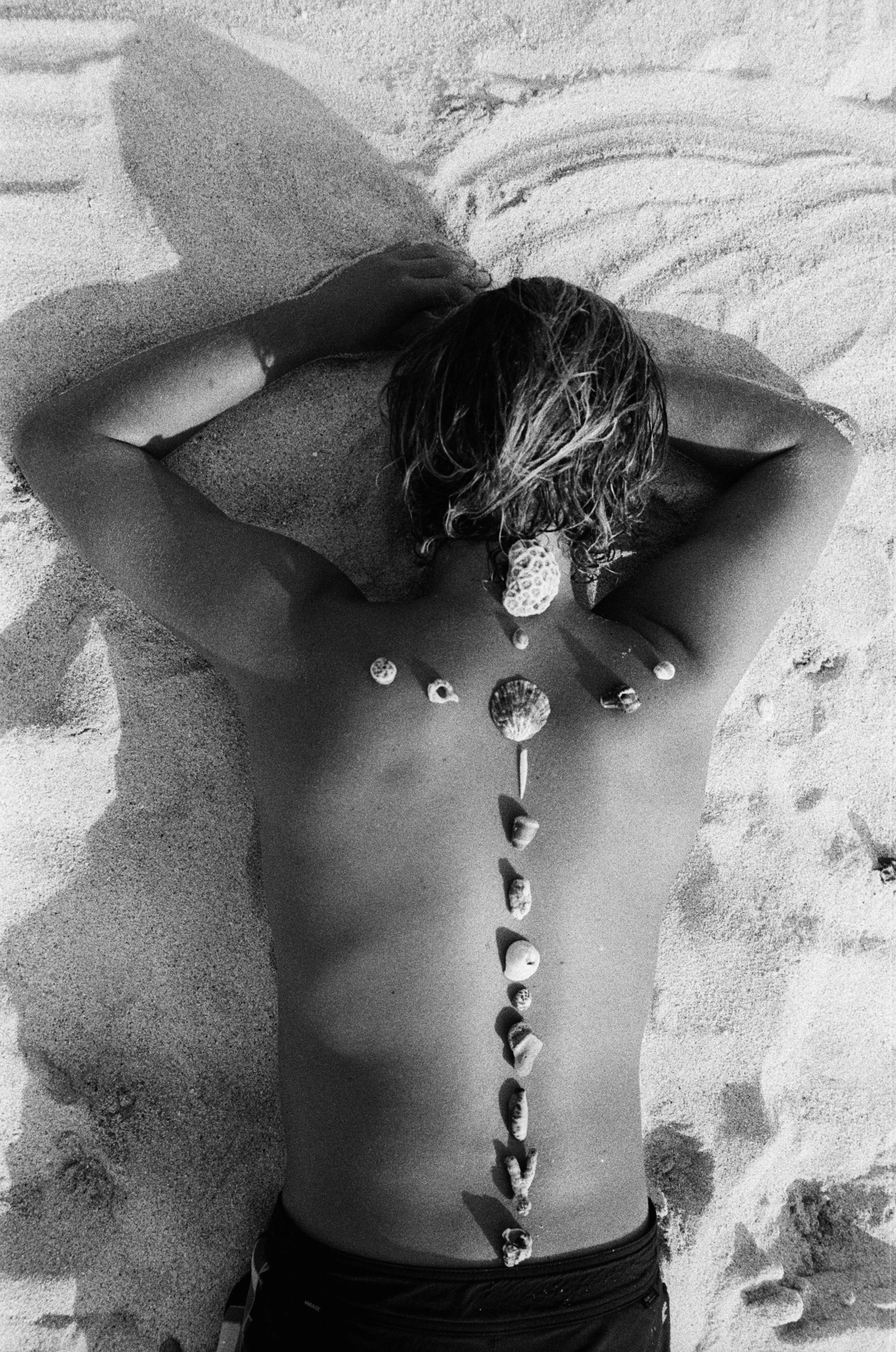 A person with wavy hair lying face down on the sand at the beach. Seashells are lined up on their back, extending from their neck to their lower back.