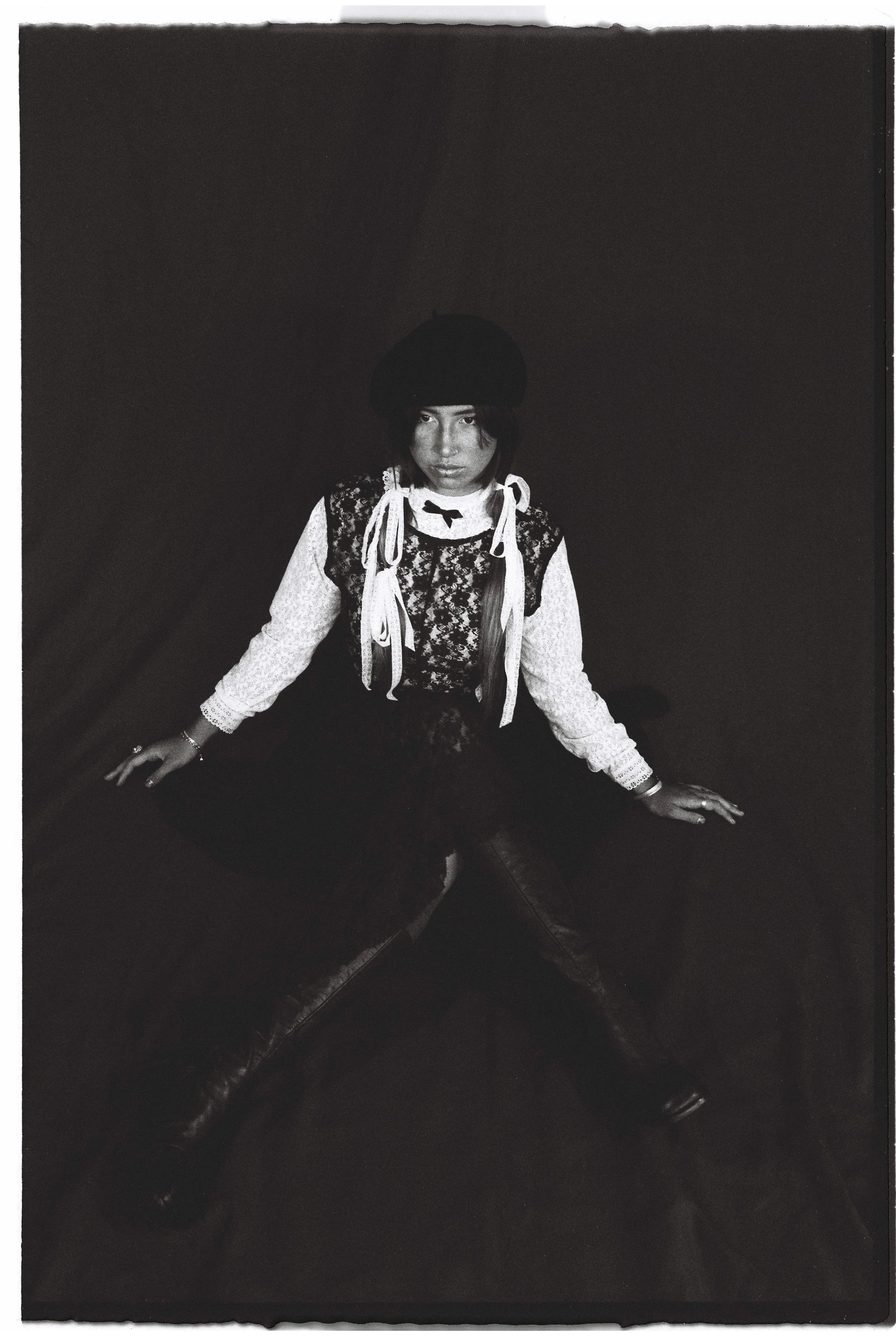 Black and white photo of a young person sitting cross-legged on a dark surface, wearing a hat, lace top, and a ribboned collar, with their hands on the surface, looking directly at the camera.