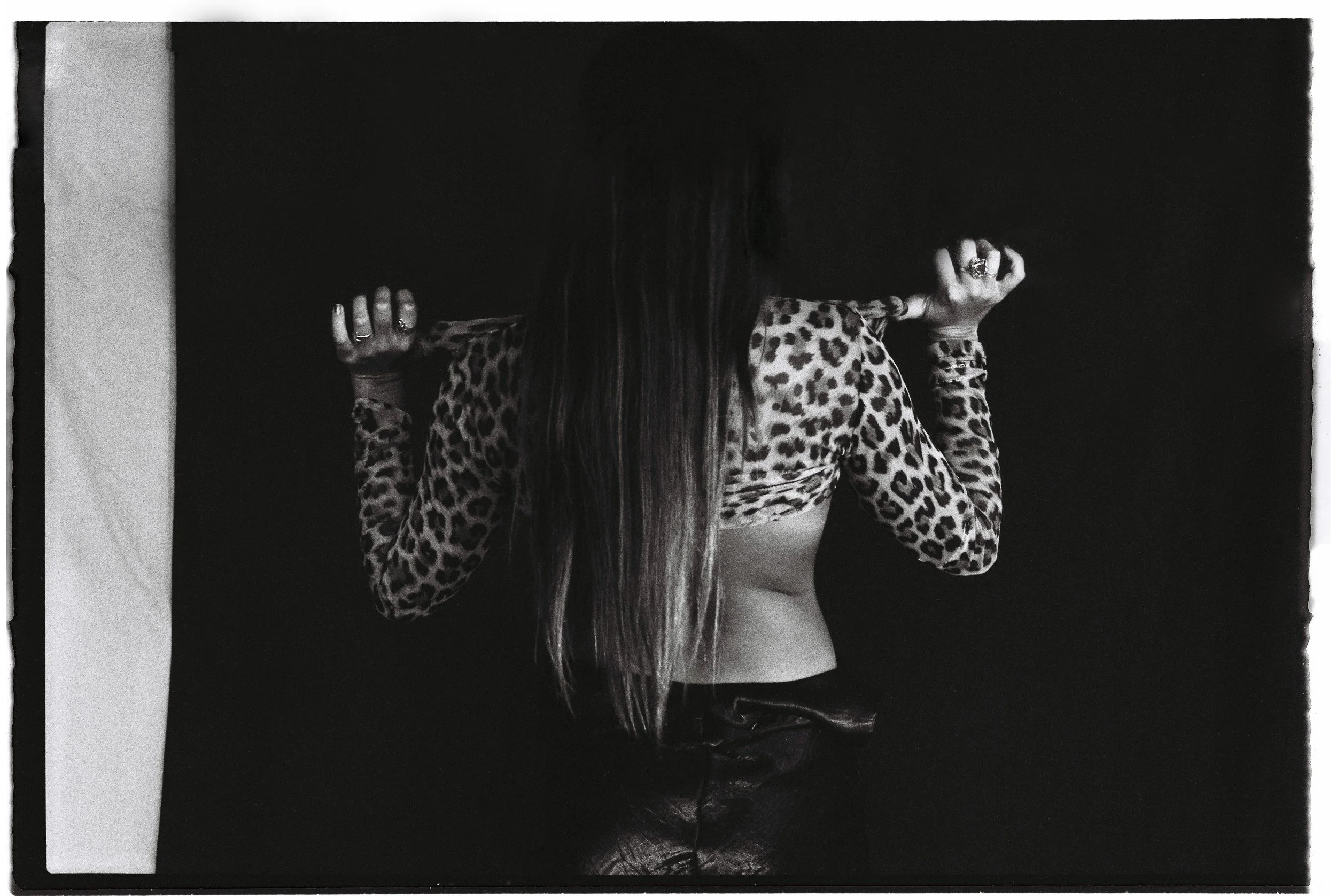 A woman with long hair holds a leopard print shirt over her shoulders, revealing her midriff, against a dark background.