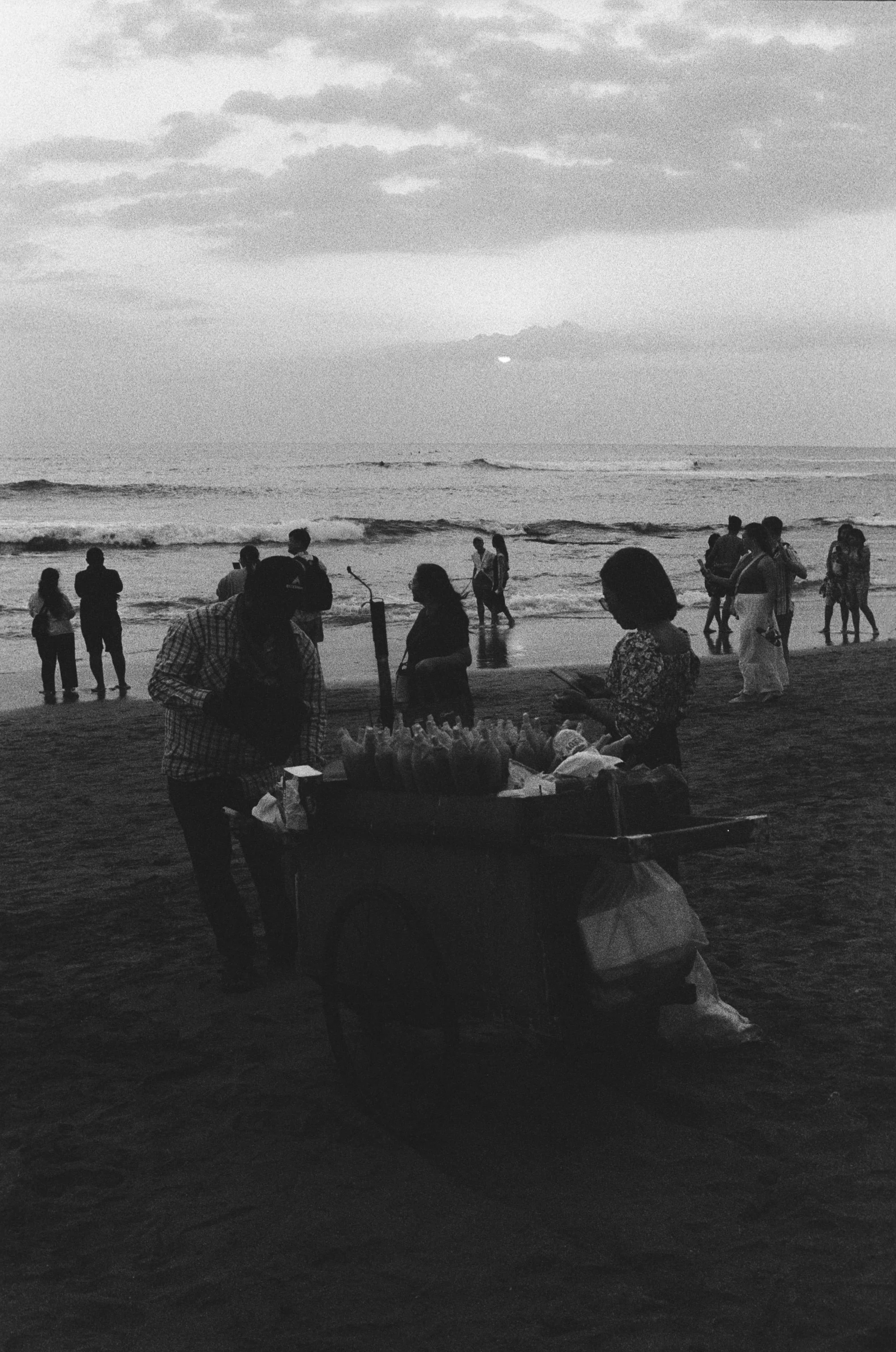 People at the beach during sunset, some are near the shore wading in the water, while others are walking or standing on the sand; there is a vendor with a cart selling snacks or drinks.