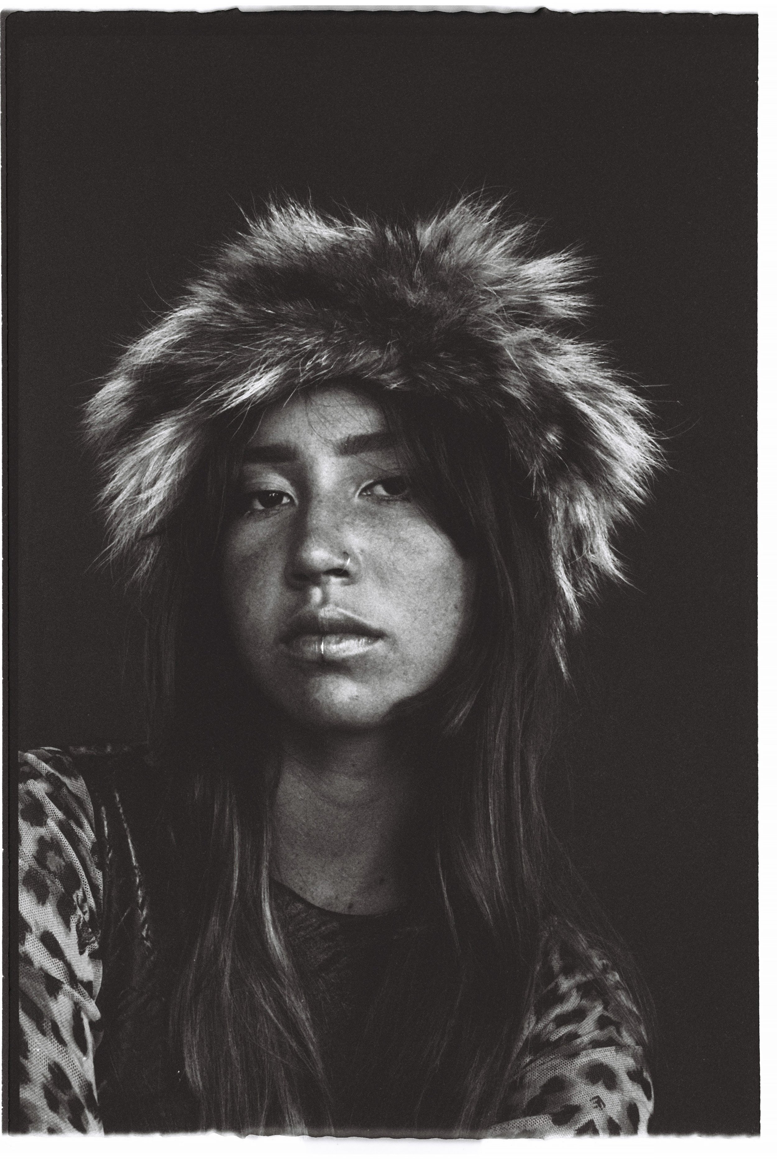 Black and white portrait of a young woman wearing a large, furry hat and a leopard print top, looking directly at the camera.