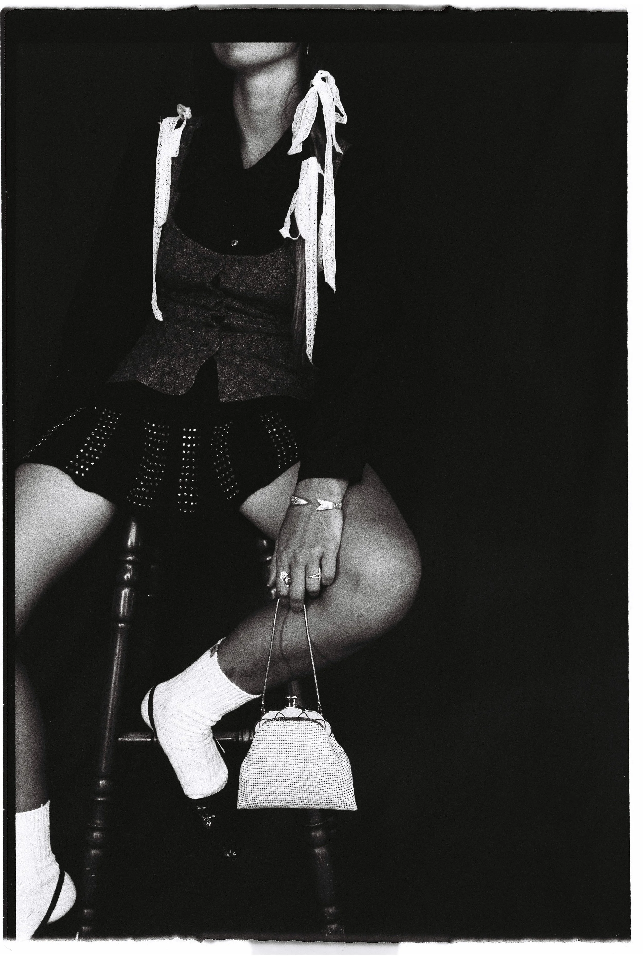 A woman sitting on a chair dressed in a patterned blazer, a black top, patterned shorts, and white socks with black shoes. She is holding a small handbag and wearing rings and bracelets.