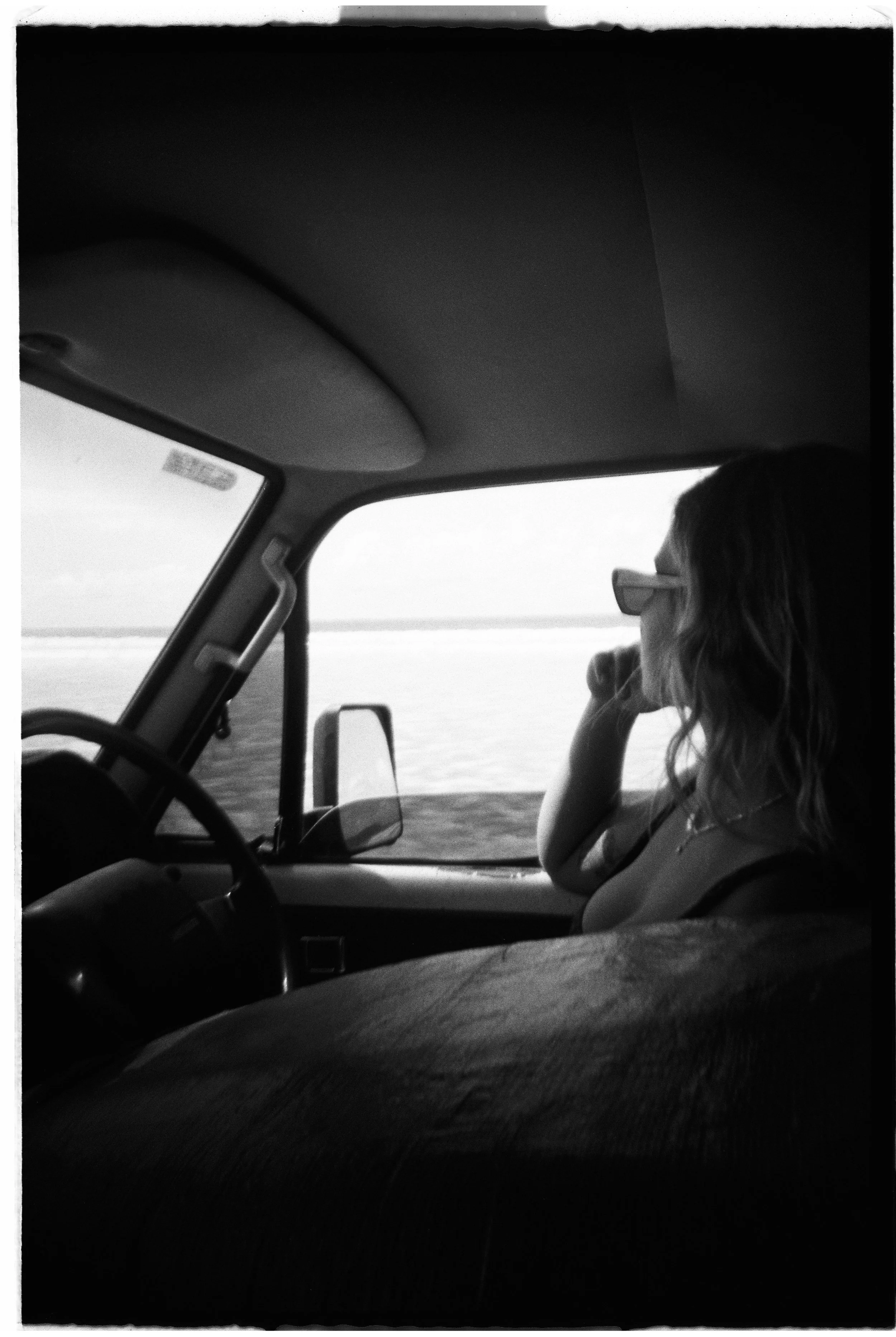 A woman with wavy hair and glasses sitting in the passenger seat of a vehicle, looking out at a body of water through the window.