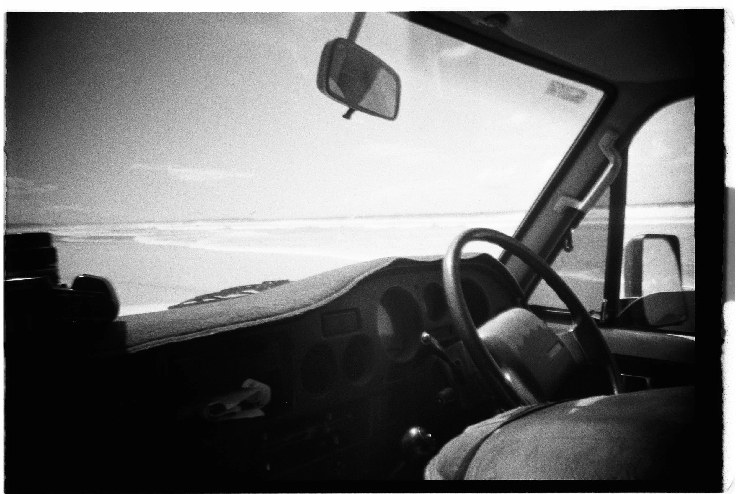 Inside view of a vehicle's dashboard and steering wheel, with a landscape of a beach and ocean visible through the windshield in black and white.