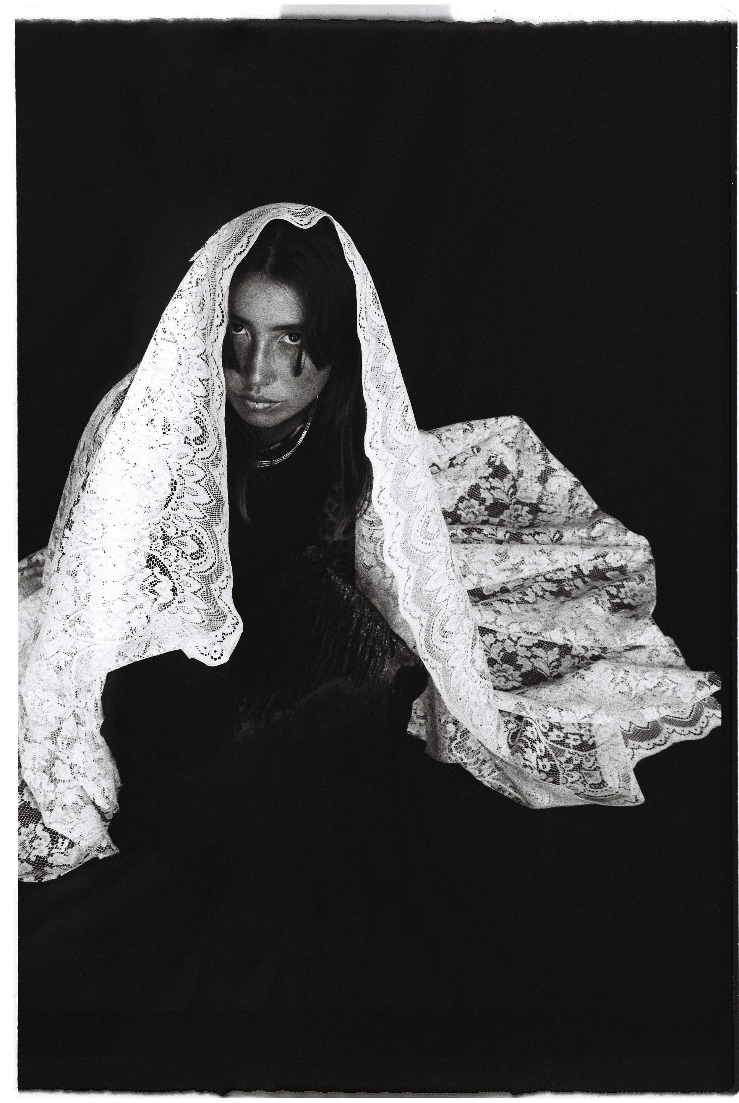 A woman with dark hair and intense eyes, partially covered by a white lace fabric, sitting on a dark surface against a black background.
