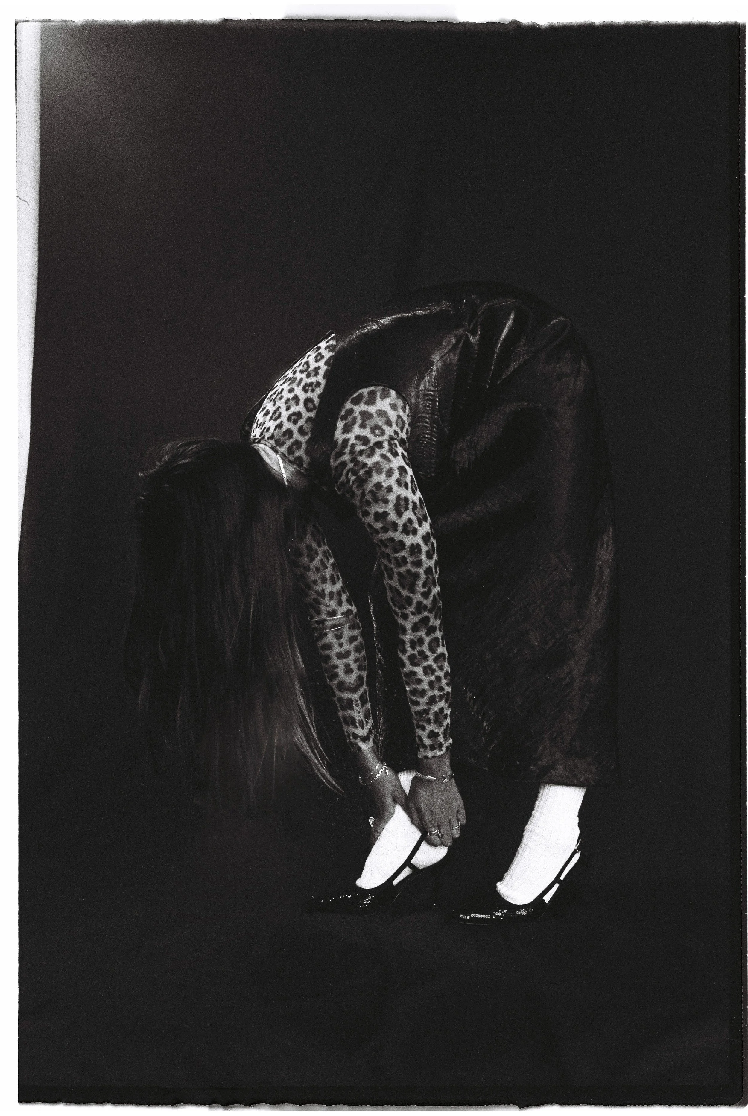 A person with long hair, dressed in leopard print top, black leather skirt, and white socks with black shoes, is bent over and adjusting or putting on their shoe against a dark background.