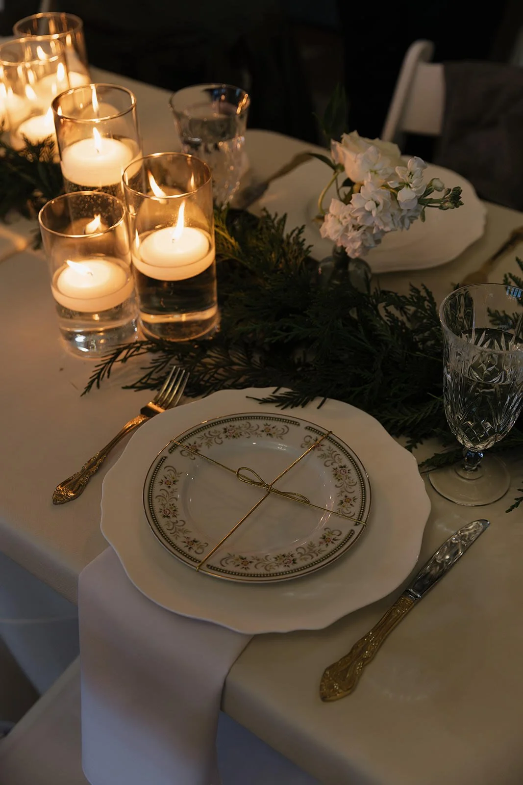 Elegant dining table with vintage plates, gold utensils, crystal glasses, a white napkin, and a floral centerpiece, illuminated by candlelight.