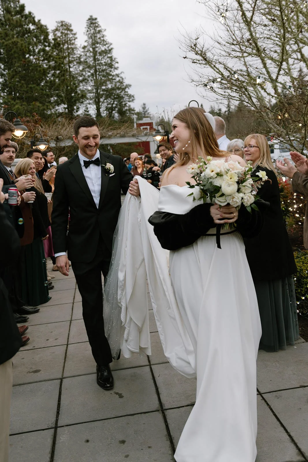 A bride holding a bouquet of white flowers walking outdoors with a groom in a black tuxedo, surrounded by smiling guests at a wedding celebration.