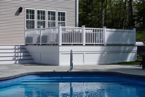 Backyard swimming pool with a deck and white fence, adjacent to a house with beige siding and multiple windows, surrounded by trees.