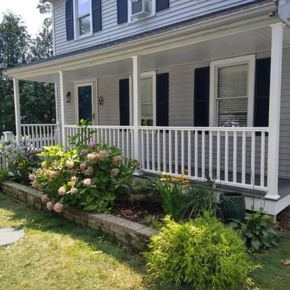 Front porch of a house with white railing, window shutters, and a garden with various plants and flowers.