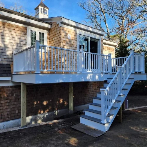 New white wooden deck with stairs attached to a house with beige shingle siding, under construction, with support posts underneath. Behind the house, there are leafless trees and a blue sky.