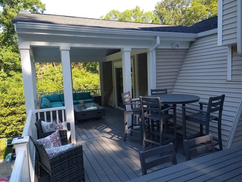 An outdoor deck with various furniture including a round black table with six chairs, a rectangular patio table with chairs, and a cushioned seating area under the porch roof. The house has beige siding and the deck is surrounded by green trees.