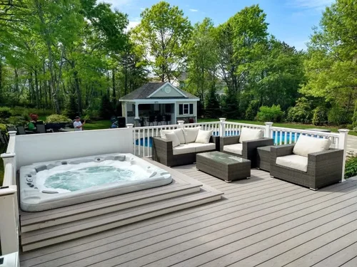 Outdoor deck area with a hot tub, wicker seating with white cushions, and a pool in the background, surrounded by trees.
