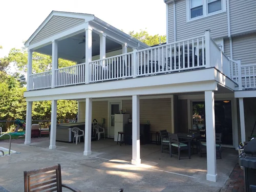 Backyard patio area with a two-story house, enclosed porch, and outdoor furniture. The house has beige siding, and the second story features a balcony with white railing. The space includes chairs, tables, and a small pool.
