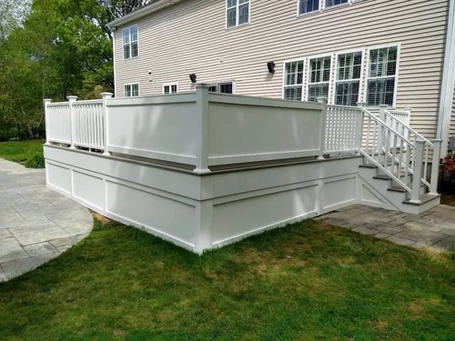A white raised deck with a railing and staircase attached to a beige house with multiple windows, surrounded by a grassy yard and some stone paving.