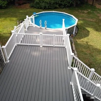 Overhead view of a backyard featuring a gray deck with white railing, leading to an above-ground swimming pool surrounded by grass and trees.