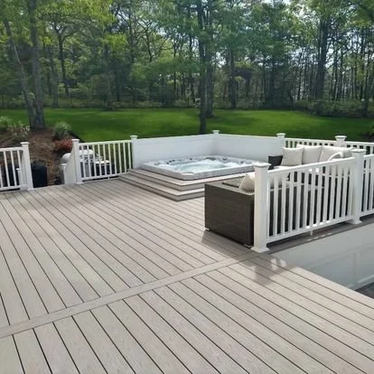 A backyard deck with a hot tub, surrounded by a white railing, and a grassy yard with trees in the background.