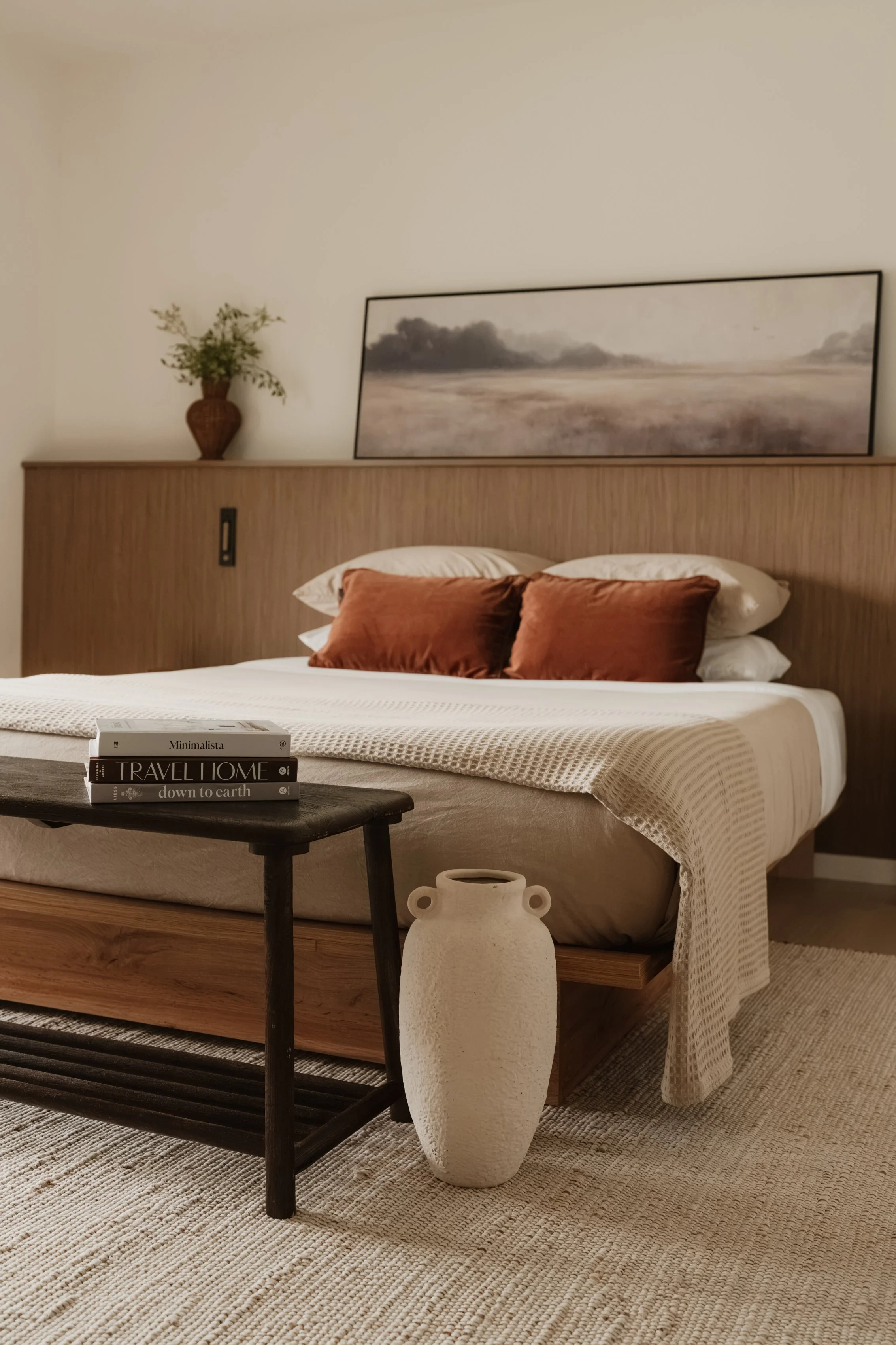 A bedroom with a bed with brown pillows, a stack of books on a black wooden tray table, a white ceramic vase on the floor, a large landscape painting, and a brass vase with greenery on the headboard.