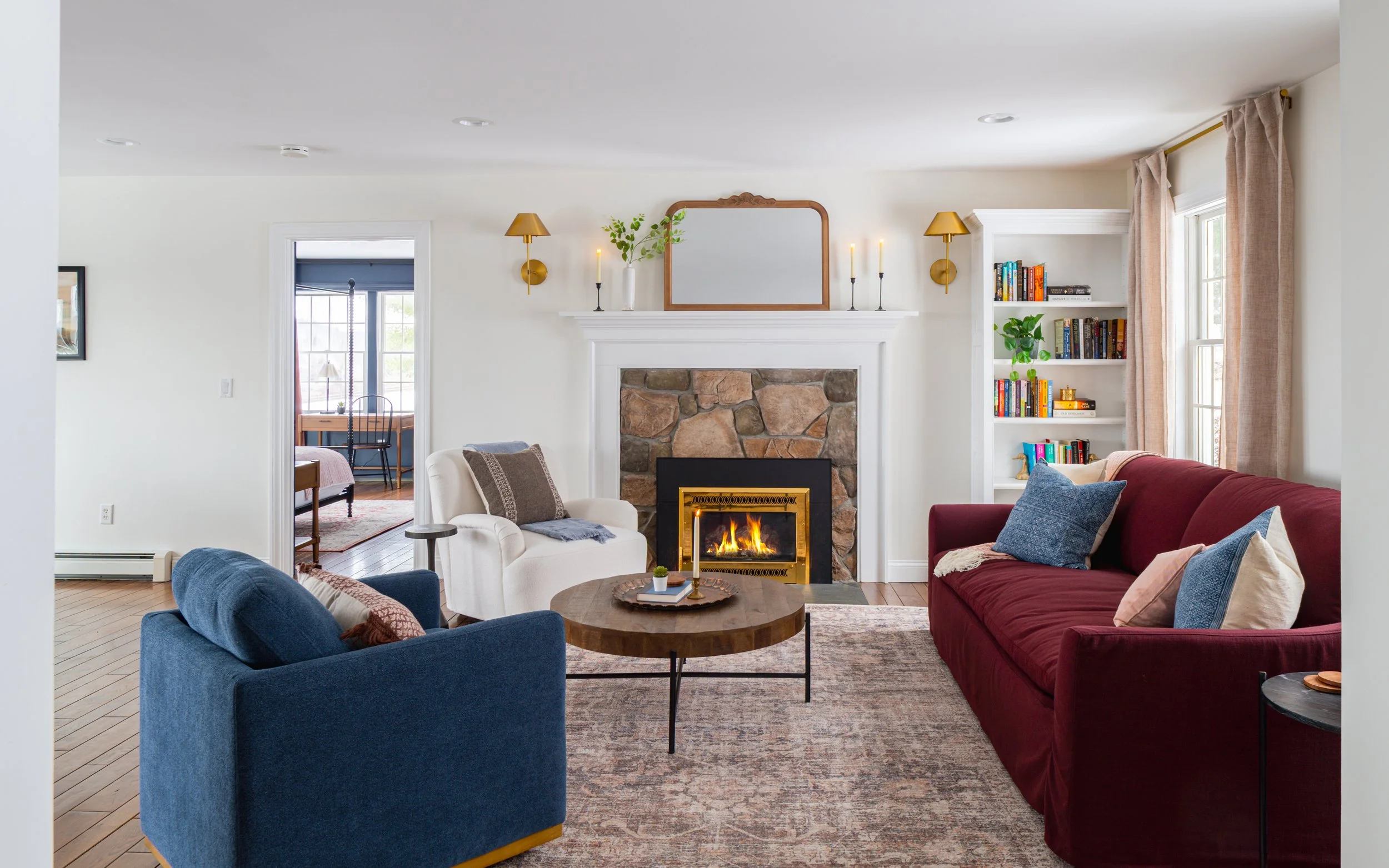 Living room with fireplace, white armchair, red sofa, blue armchair, wooden coffee table, shelves with books, and decor, decorated for cozy ambiance.