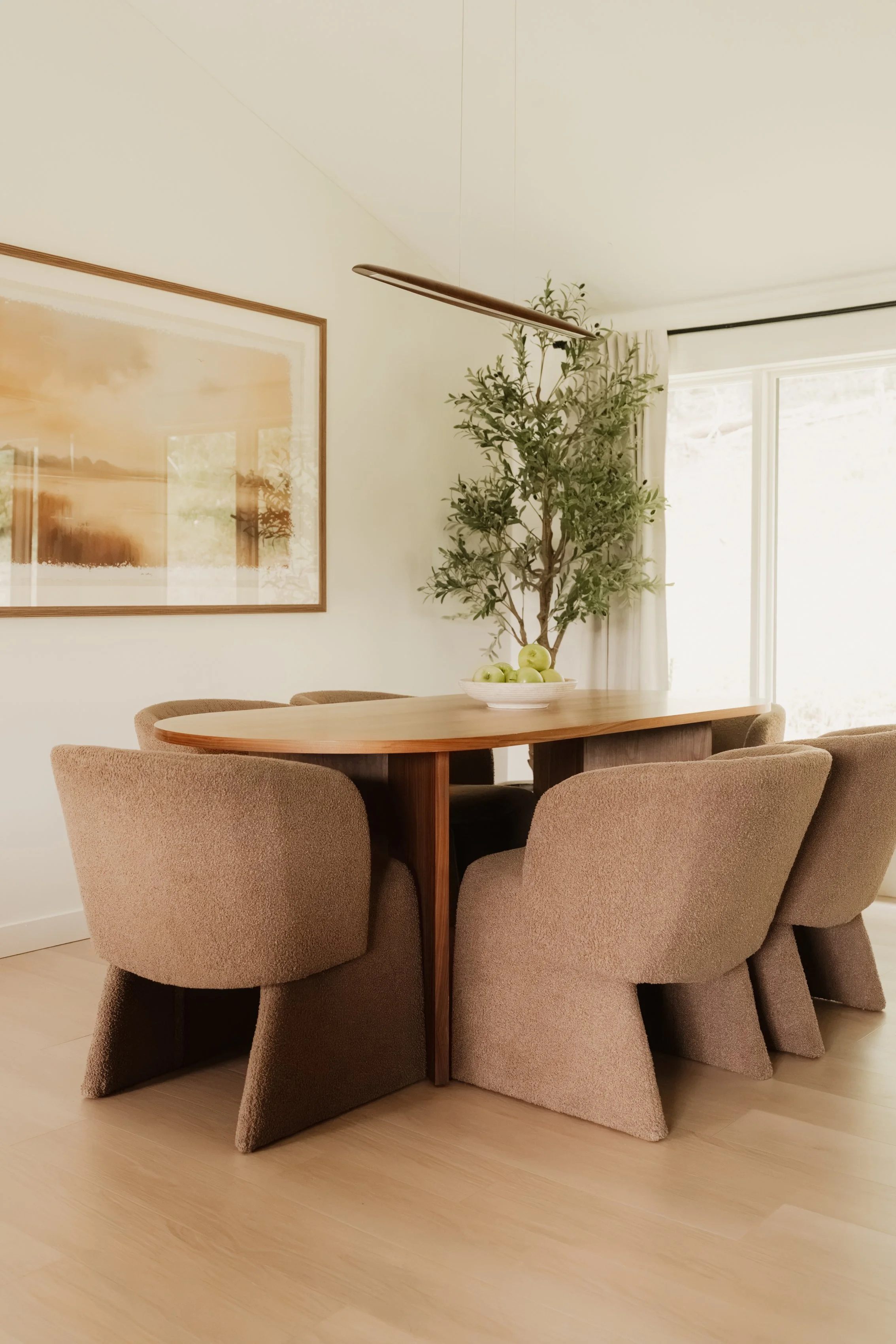 A dining room with a wooden table surrounded by six beige upholstered chairs. A bowl of green apples is on the table. There is a large leafy plant behind the table and a framed artwork on the wall. A window with white curtains lets in natural light.