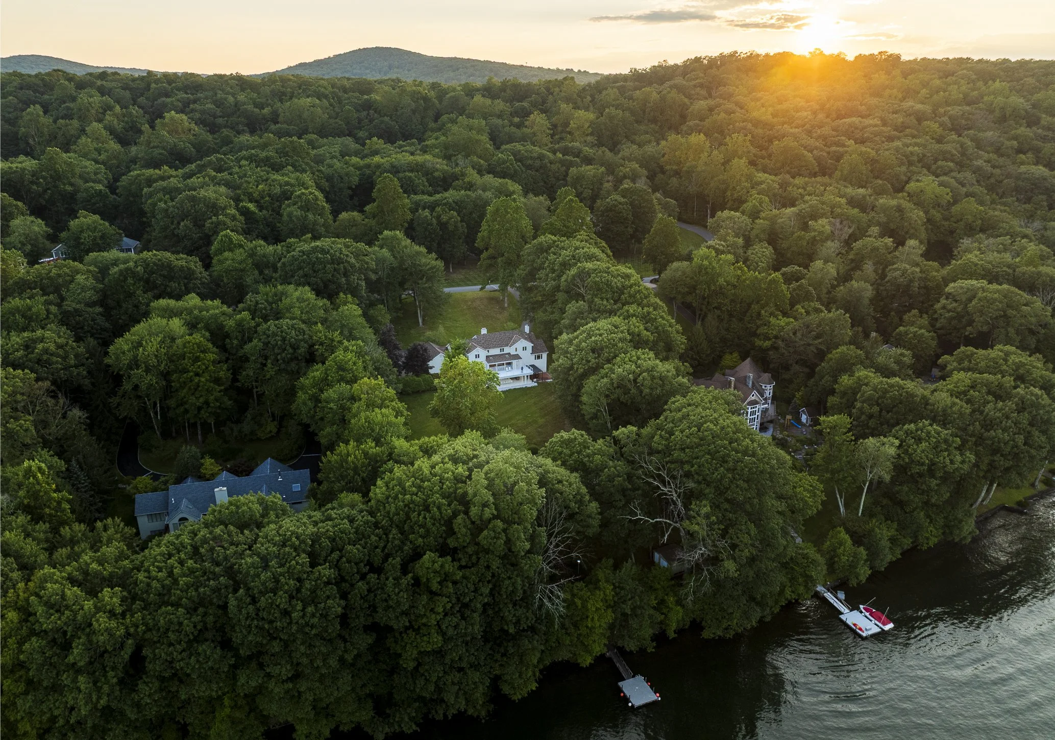Aerial view of a lakeside area with lush green trees, houses with boats docked at piers, and hills in the background at sunset.