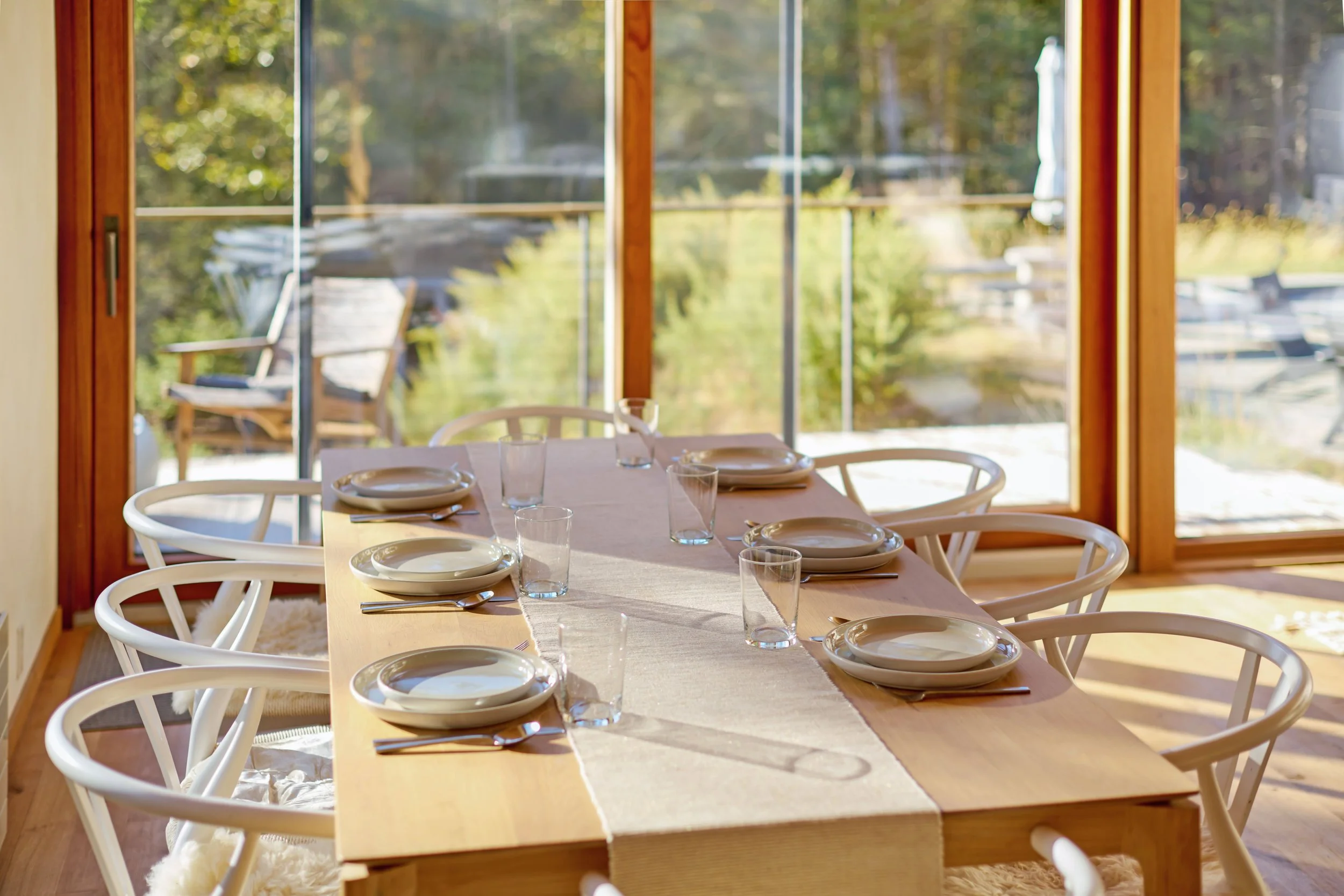 Dining table set with plates, glasses, and silverware in front of large windows overlooking a deck and trees outside.