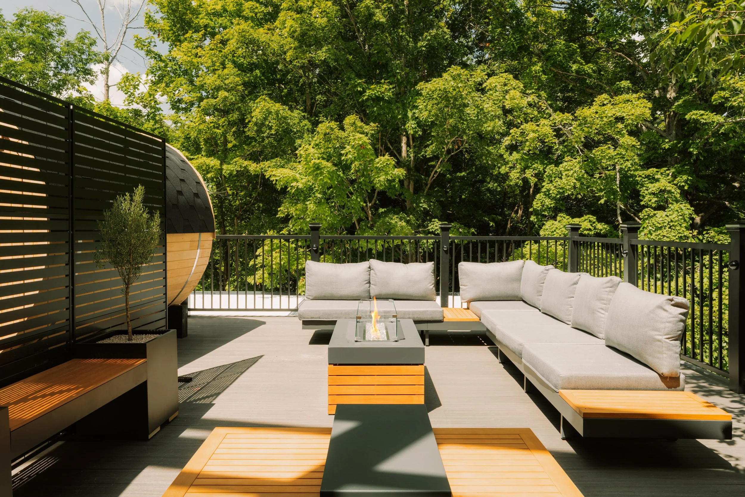 Modern outdoor patio with beige cushioned sectional sofa, a fire pit, wooden side tables, and a privacy screen, surrounded by lush green trees.